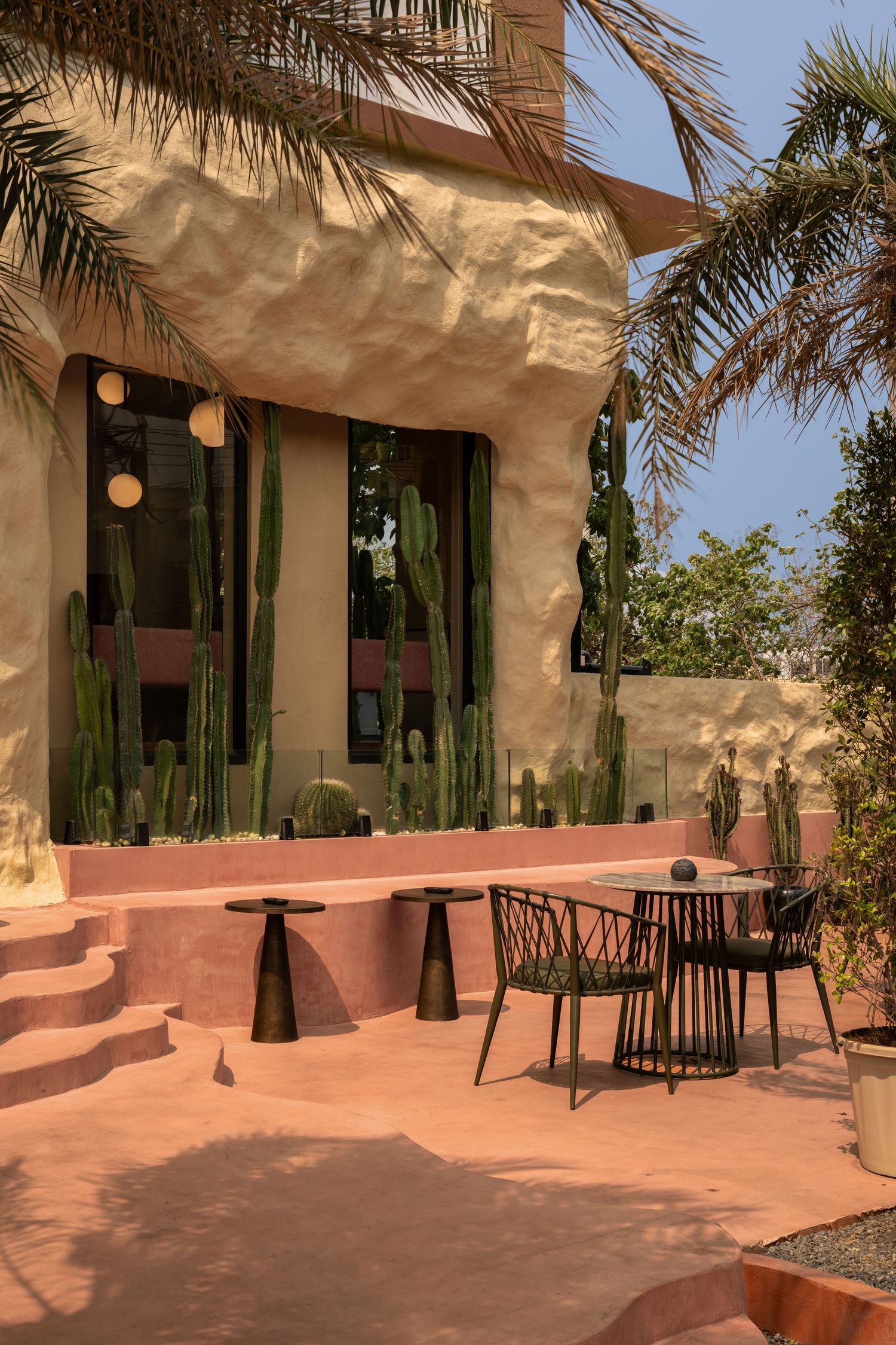 Desert-inspired restaurant exterior with pink terrace, cacti, palm trees, and textured sandy walls.