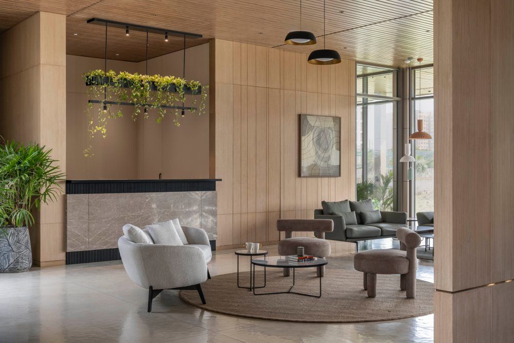 Modern hotel lobby with warm wood panels, hanging greenery, white curved chairs, and pendant lights over a circular rug.