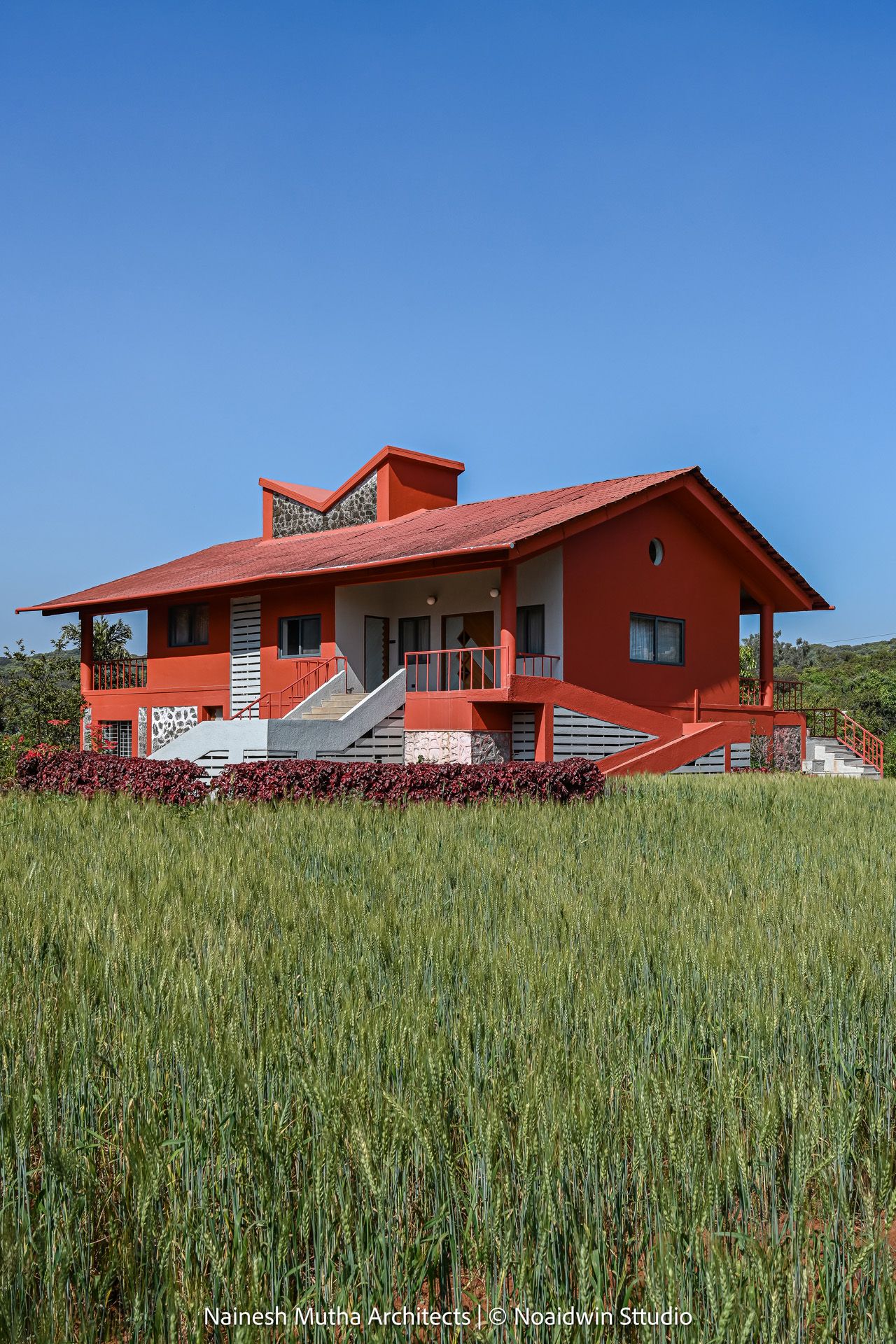 Striking red farmhouse with gabled roof set amid green crops under clear blue sky.