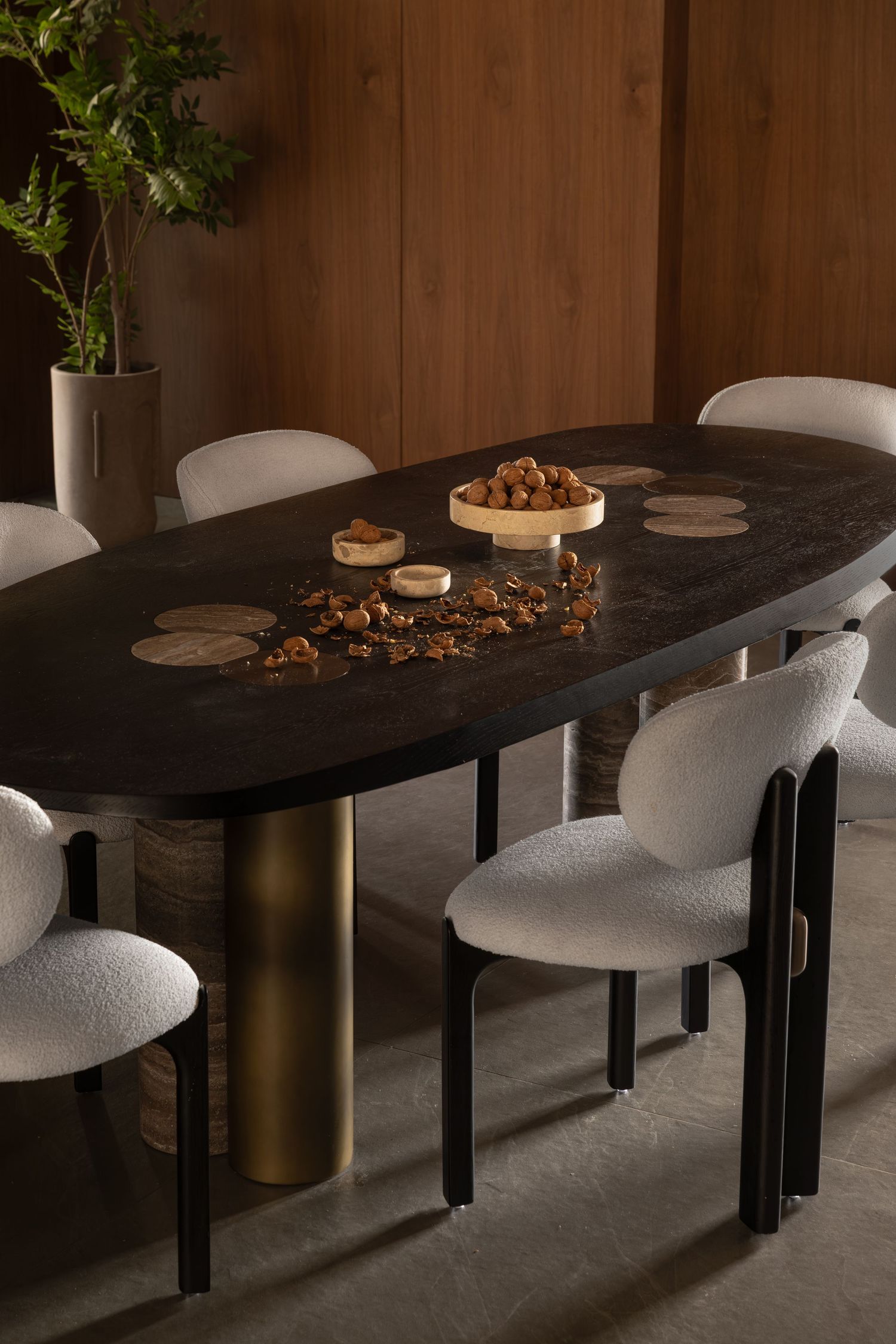 Dark oval dining table with brass legs, white bouclé chairs, decorative bowls, and warm wood panel backdrop.