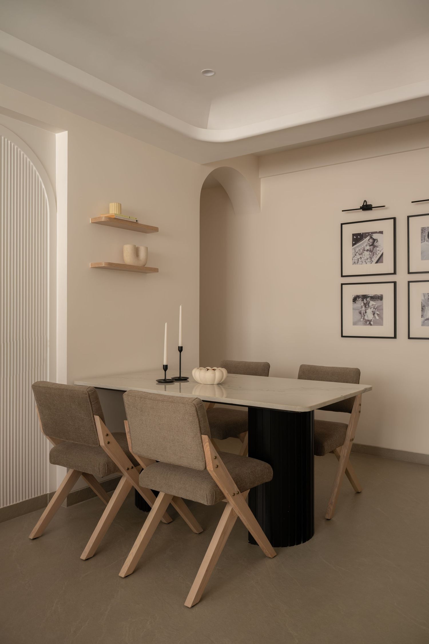 Minimalist dining room with beige upholstered chairs, marble-top table, black candleholder, floating shelves, and framed art on walls.