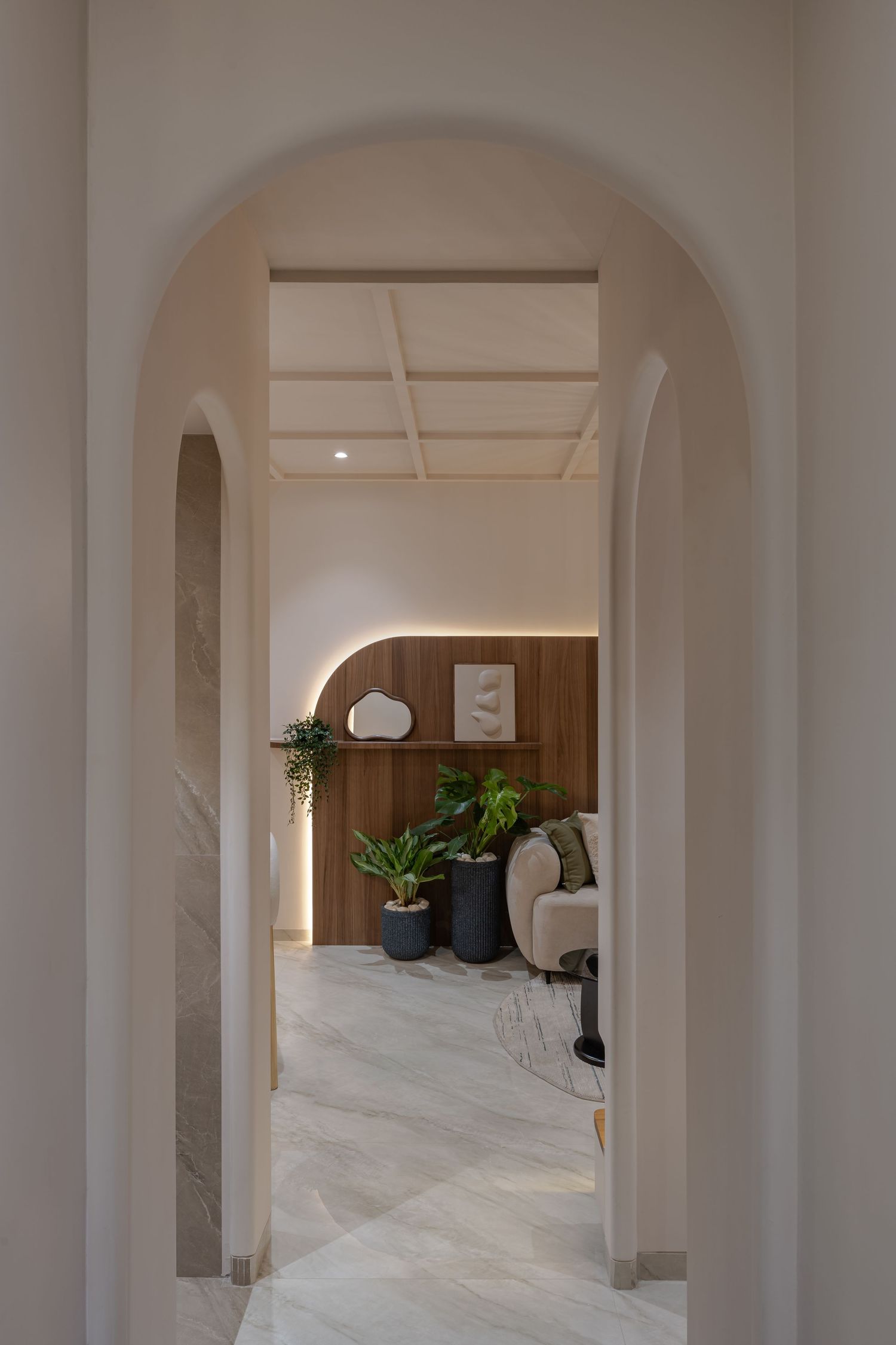 Elegant arched corridor in neutral tones leading to a cozy sitting area with tropical plants and wood paneling.