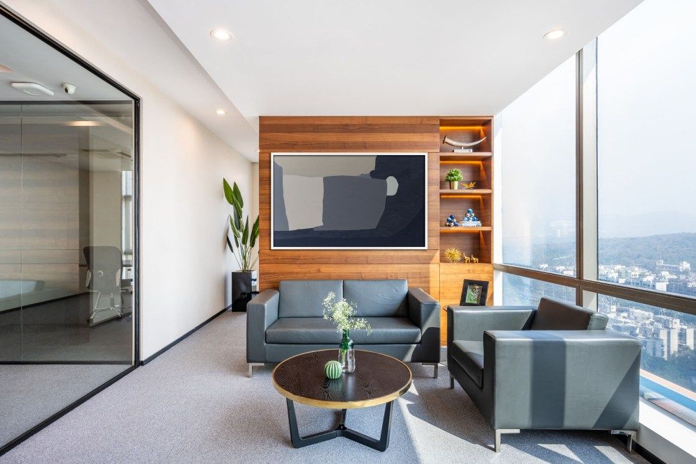 Contemporary office lounge with gray sofas, wooden built-in shelving, wall-mounted TV, and panoramic city views.
