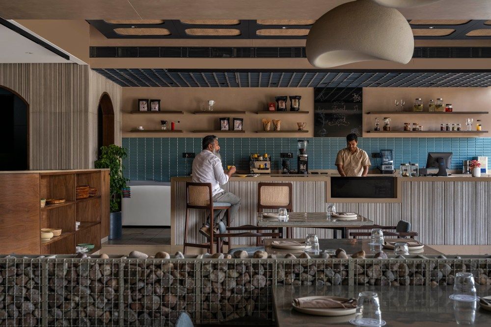 Restaurant bar area with blue tile backsplash, open shelving, rattan bar stools, and gabion stone partition in foreground.