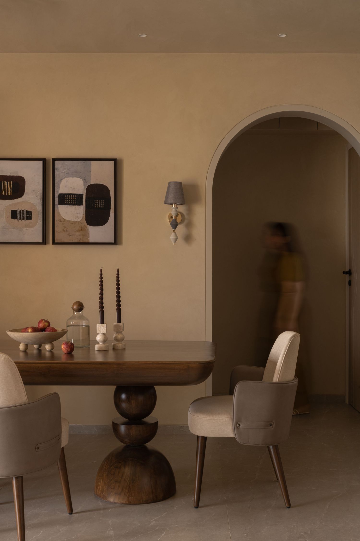 Warm-toned dining room with sculptural wooden table, cream chairs, abstract art, and arched doorway.