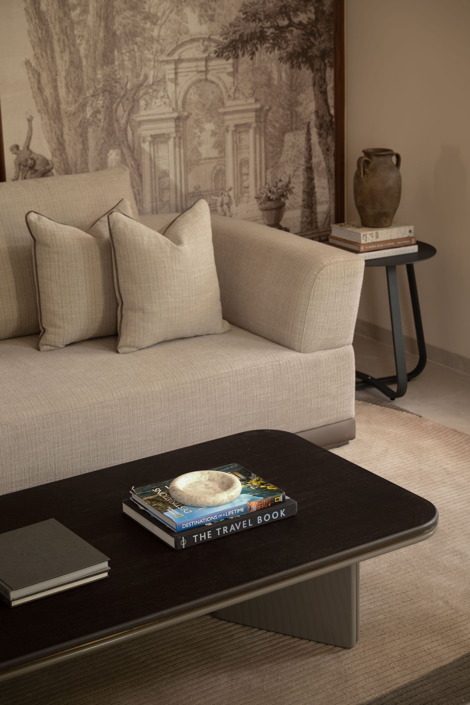 Detail shot of beige sofa with cushions, dark coffee table with books, and classical landscape mural.