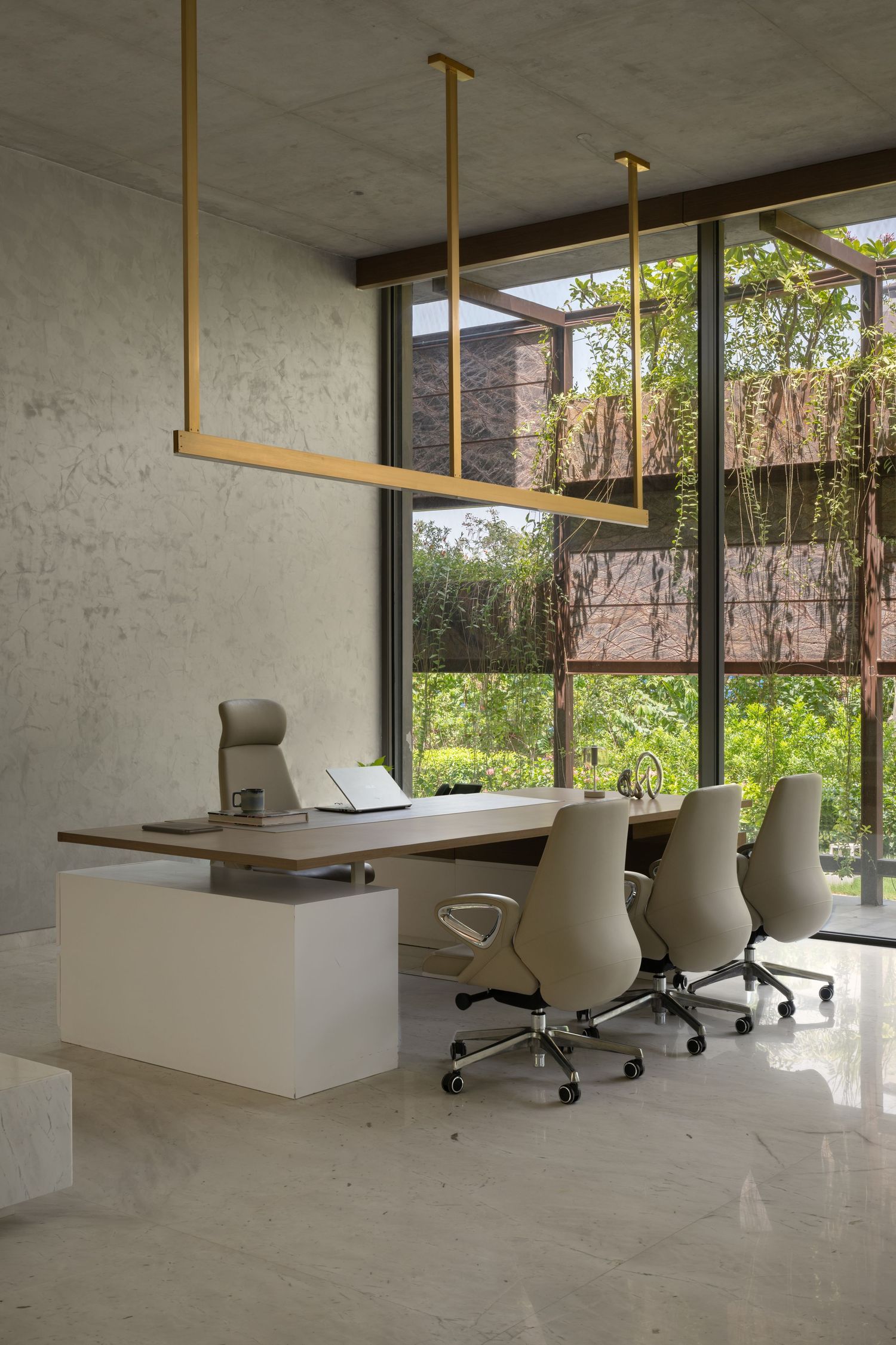 Modern office workspace with gold pendant lights, beige ergonomic chairs, and floor-to-ceiling windows overlooking lush greenery.