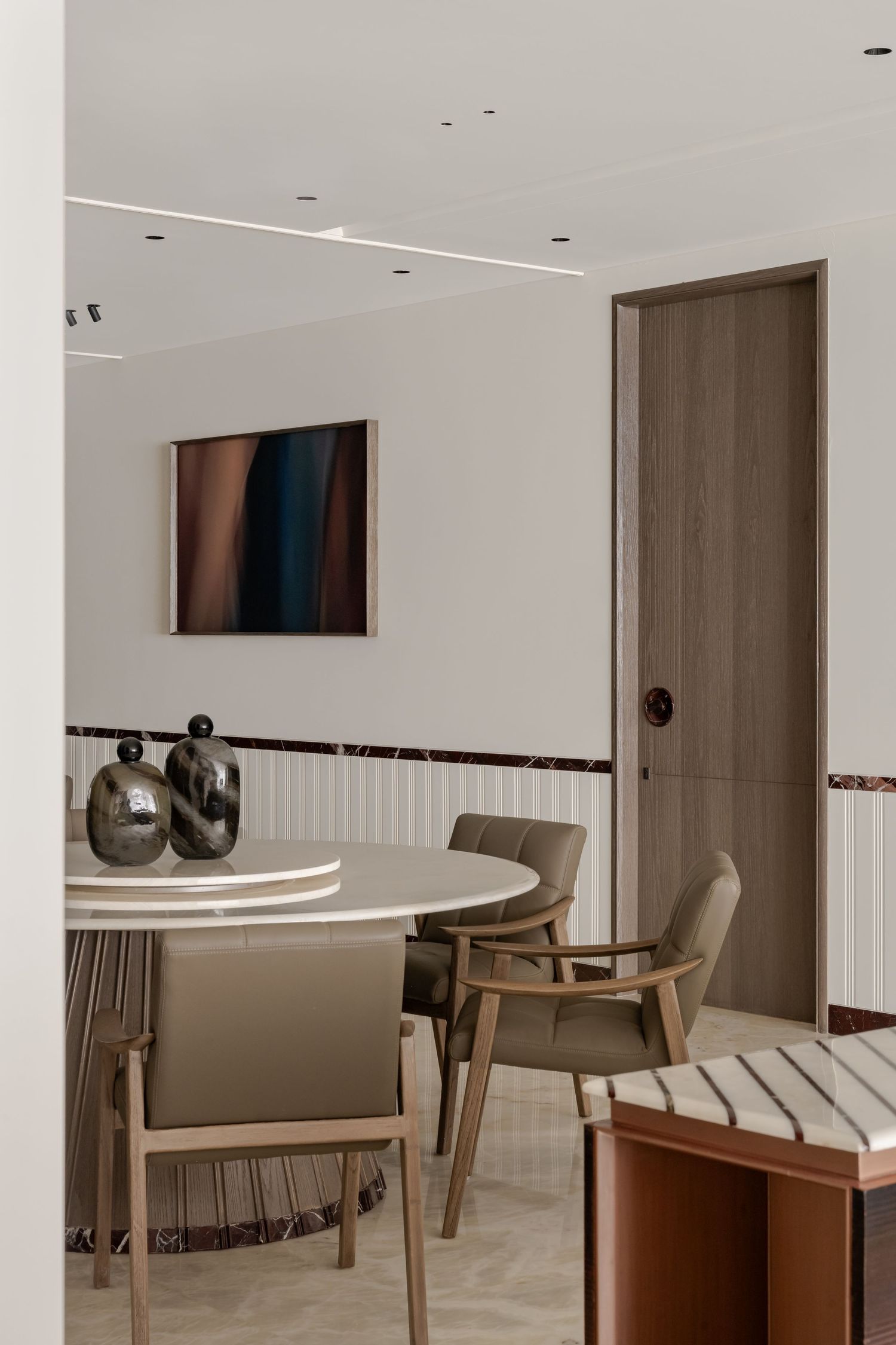 Elegant dining room with round white table, taupe chairs, metallic vases, wood door, and abstract artwork on white walls.