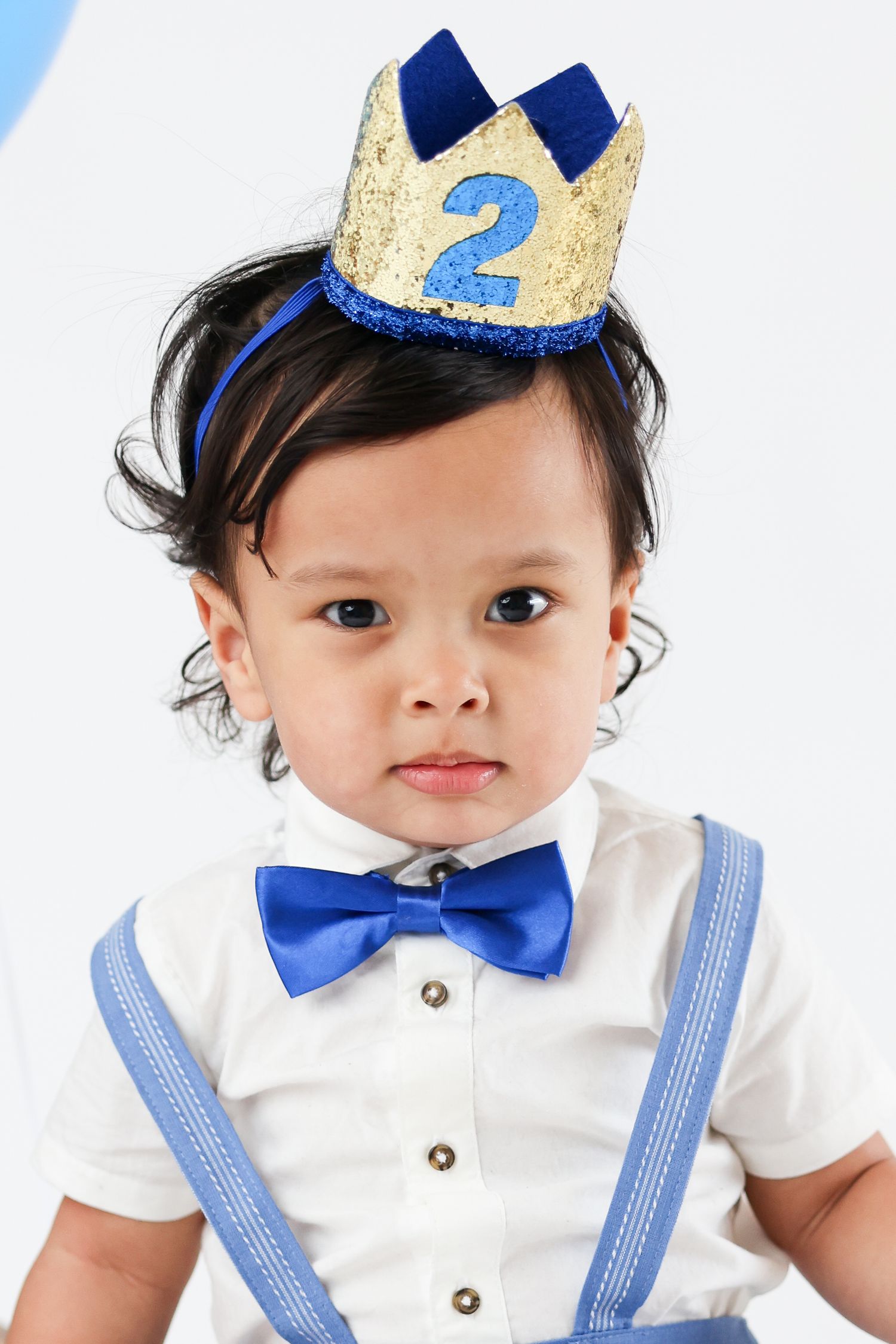 Toddler boy wearing a gold crown with number 2, blue bow tie, and suspenders celebrating his 2nd birthday.