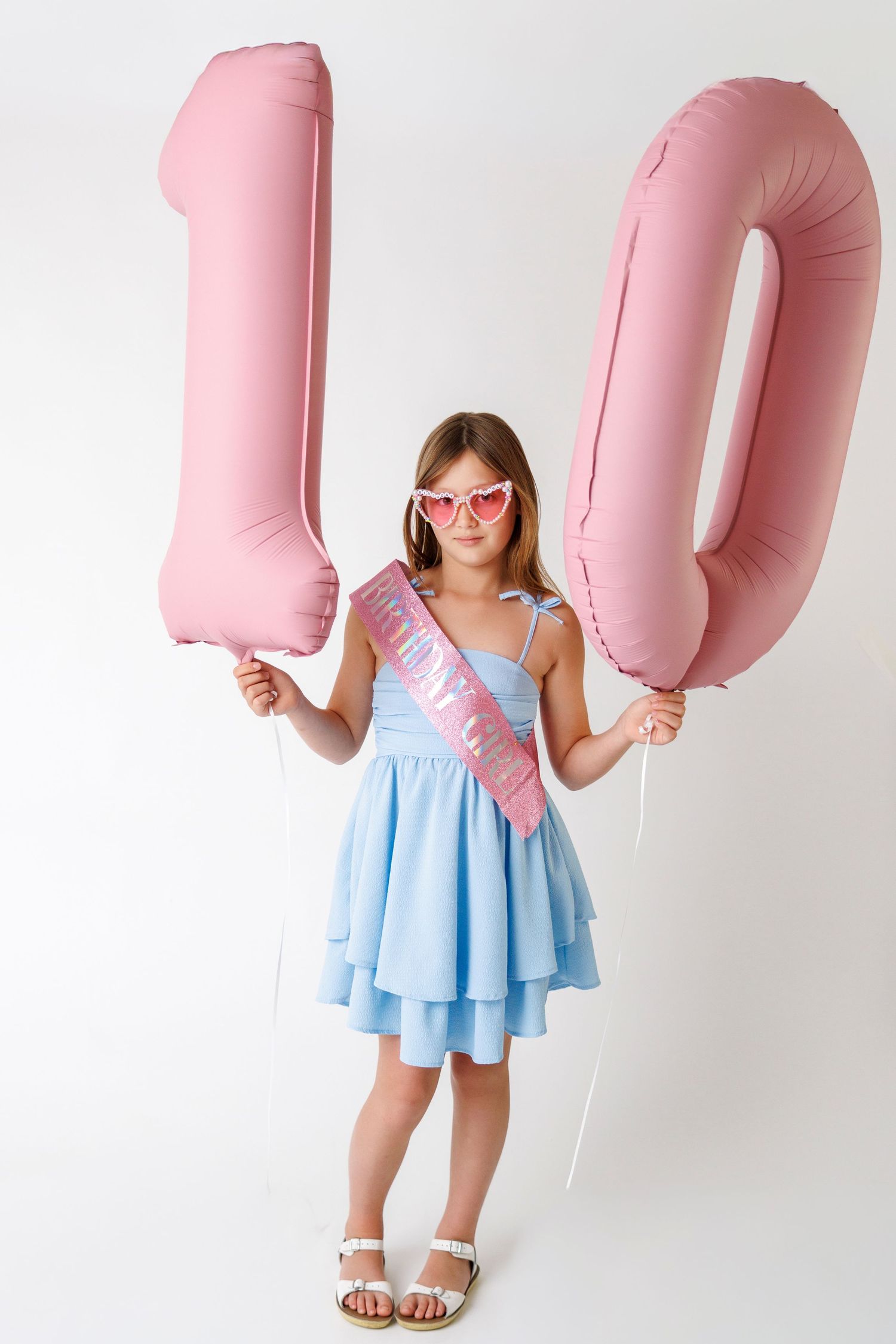 A girl in a blue dress holds pink number 10 balloons, wearing heart sunglasses and a birthday sash.
