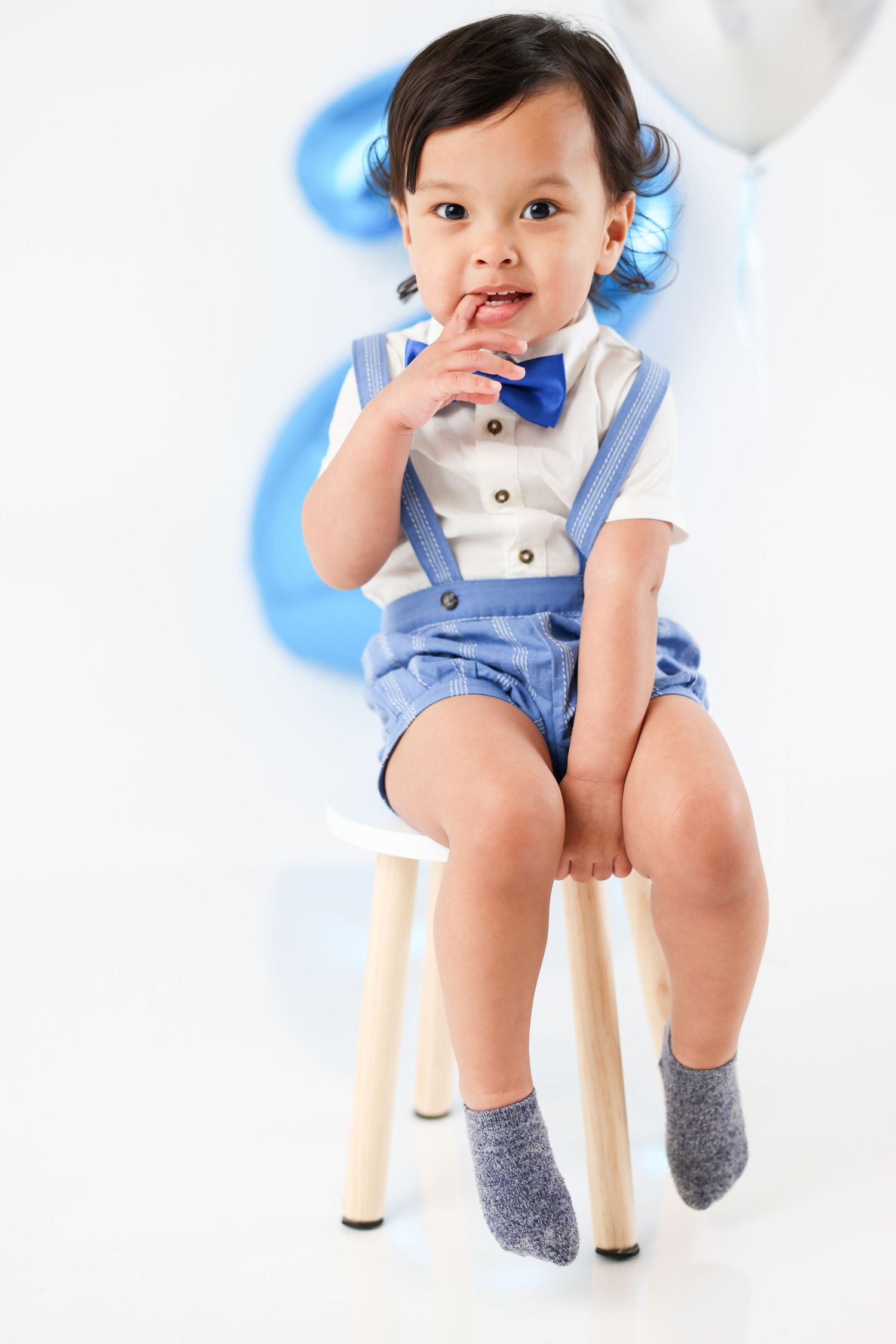 Adorable baby boy in blue suspender shorts and bow tie sits on small chair with blue balloons backdrop.