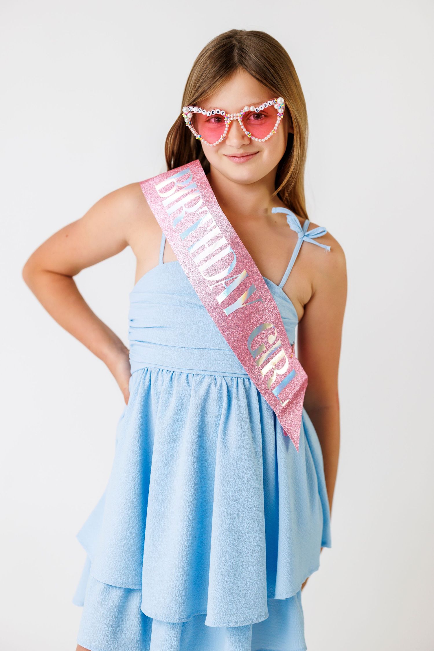 Young girl in blue dress wearing a pink glittery Birthday Girl sash and heart-shaped sunglasses posing confidently.
