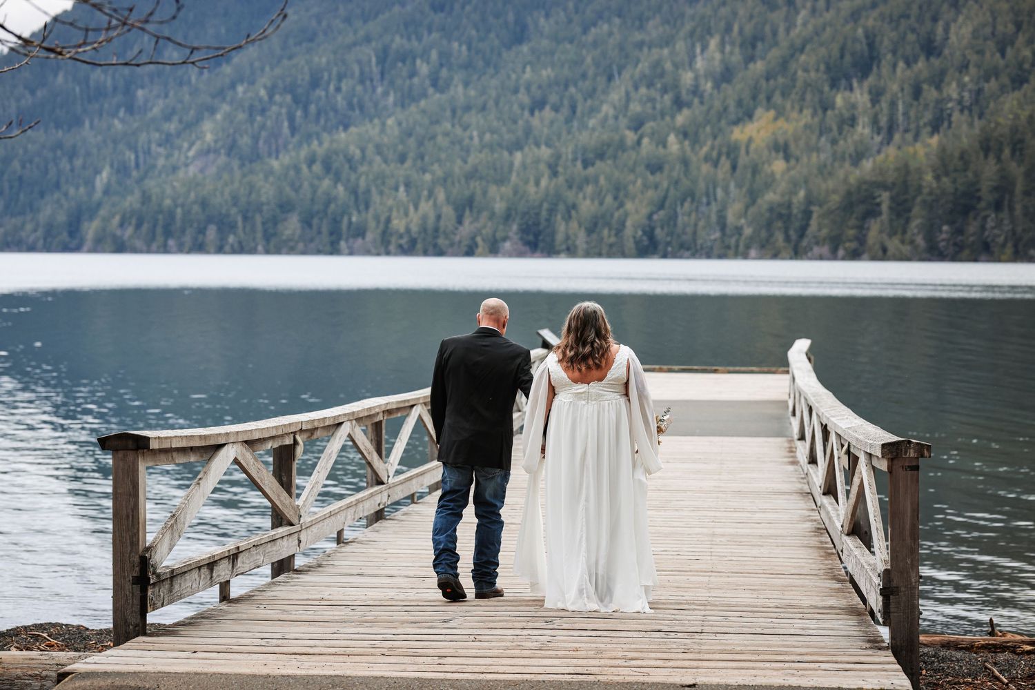 A couple stands together on a wooden dock overlooking a serene mountain lake surrounded by evergreen forest.