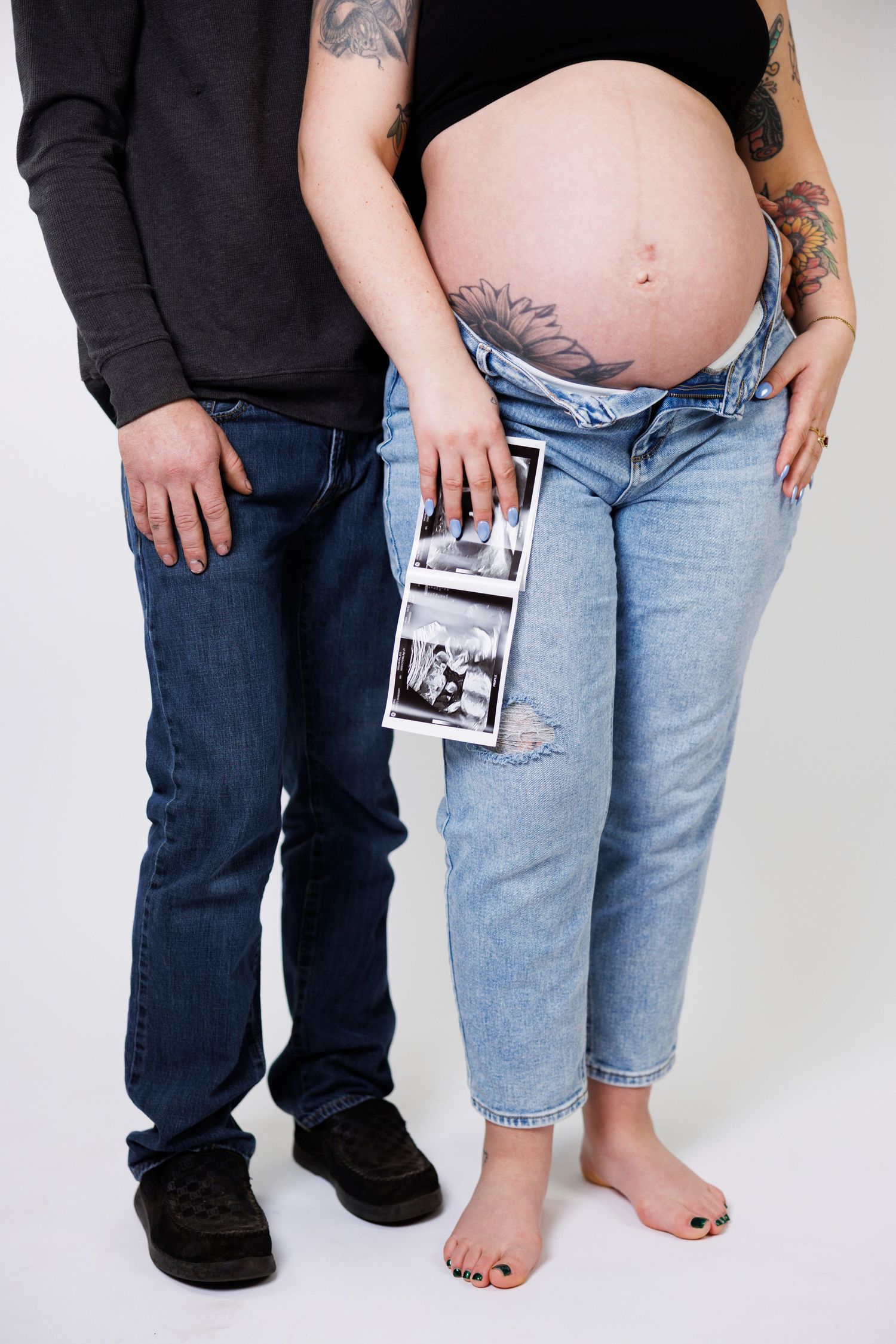 Couple standing together showing pregnancy belly while holding ultrasound photo, wearing casual denim and black tops.