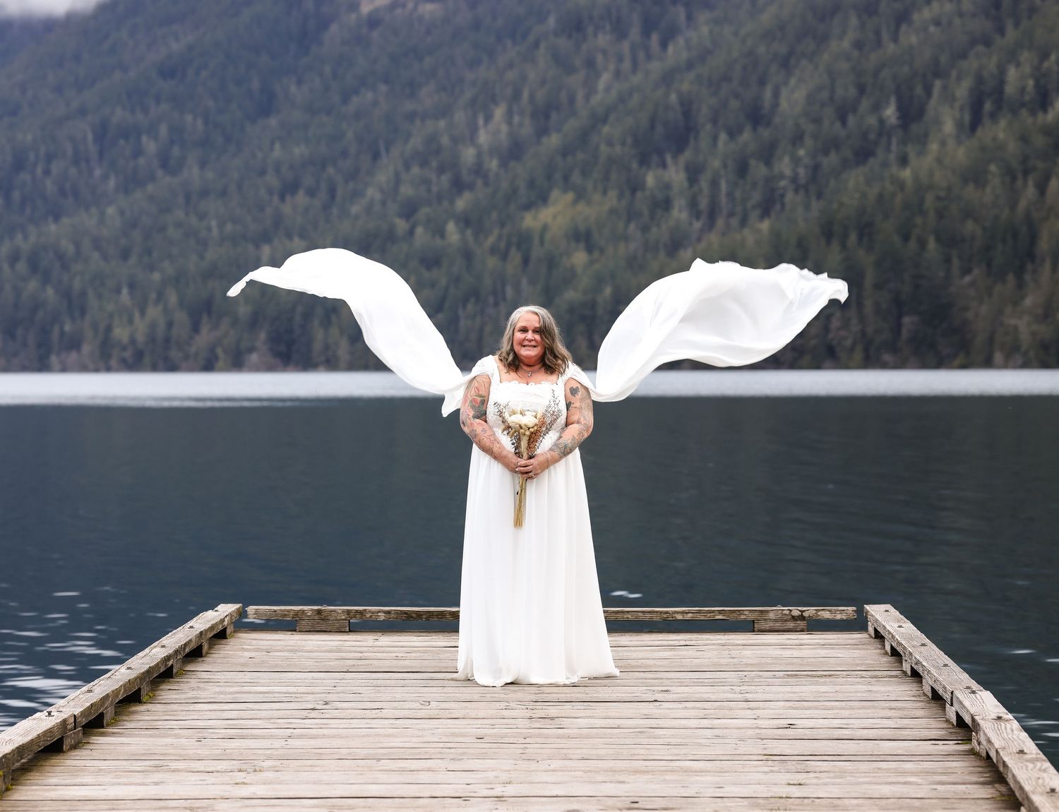 A bride in flowing white dress poses on a wooden dock with mountains and lake in background.