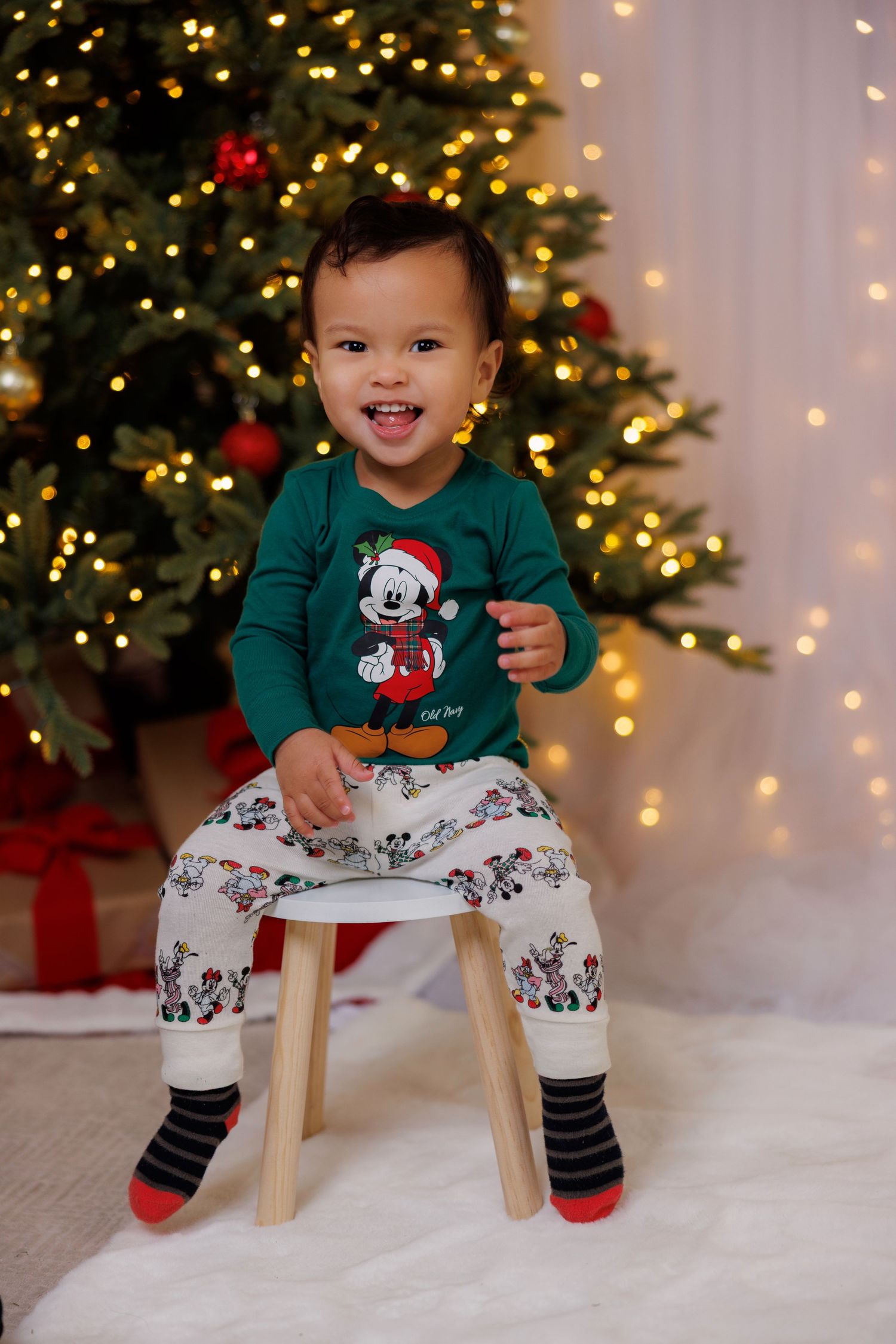 Festive holiday scene with child wearing Mickey Mouse Christmas sweater sitting on stool by decorated tree and lights.