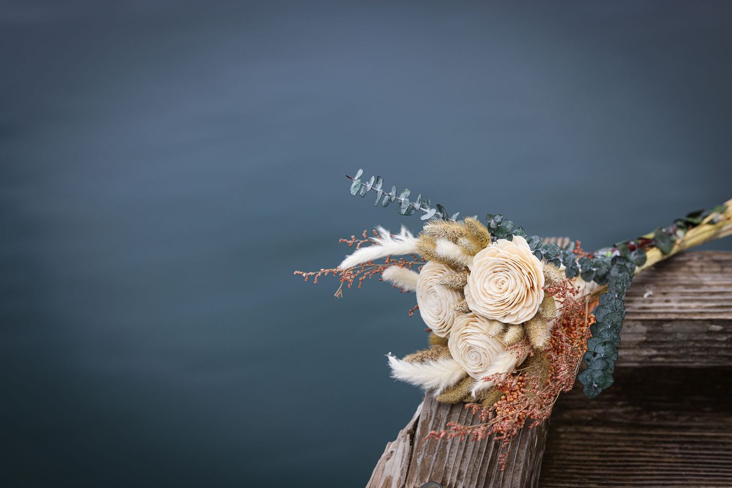 Rustic wedding bouquet with cream roses and dried flowers rests on wooden dock by a lake.