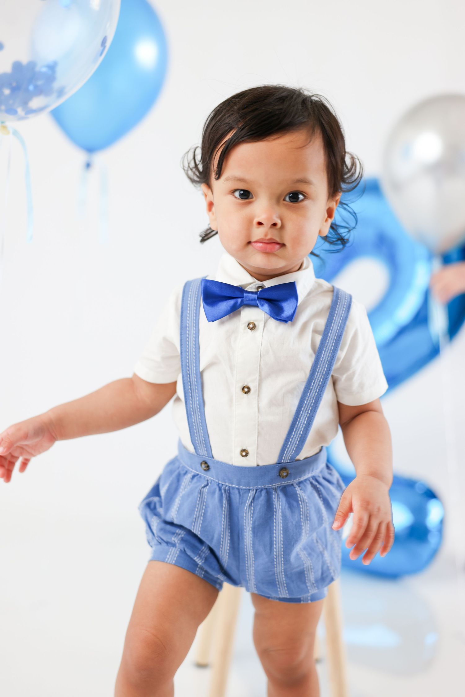 Adorable baby boy in white shirt, blue bow tie, suspenders, and shorts poses for first birthday photos with blue balloons.
