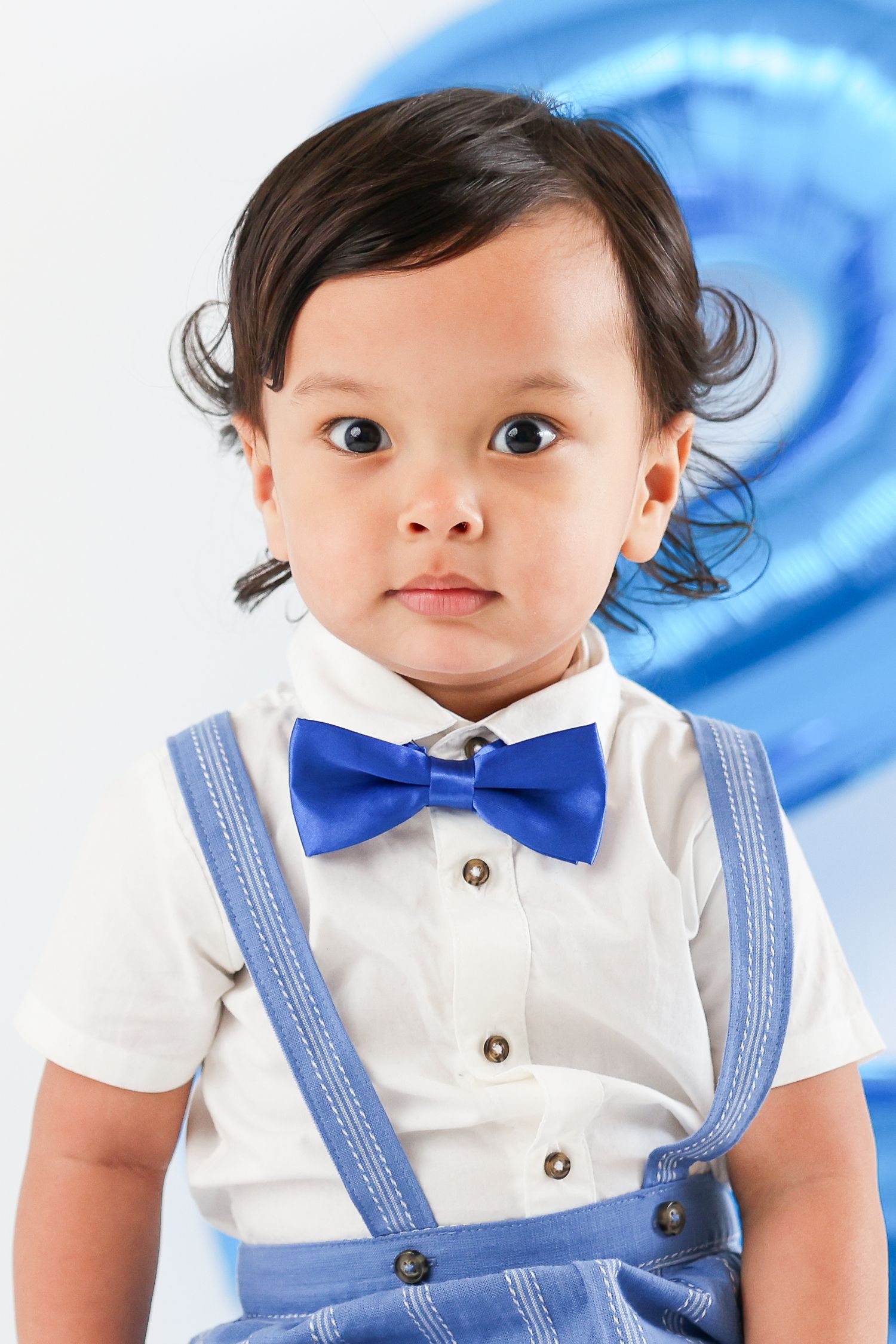 Adorable toddler boy wearing blue bow tie suspenders outfit poses in front of blue number balloon.
