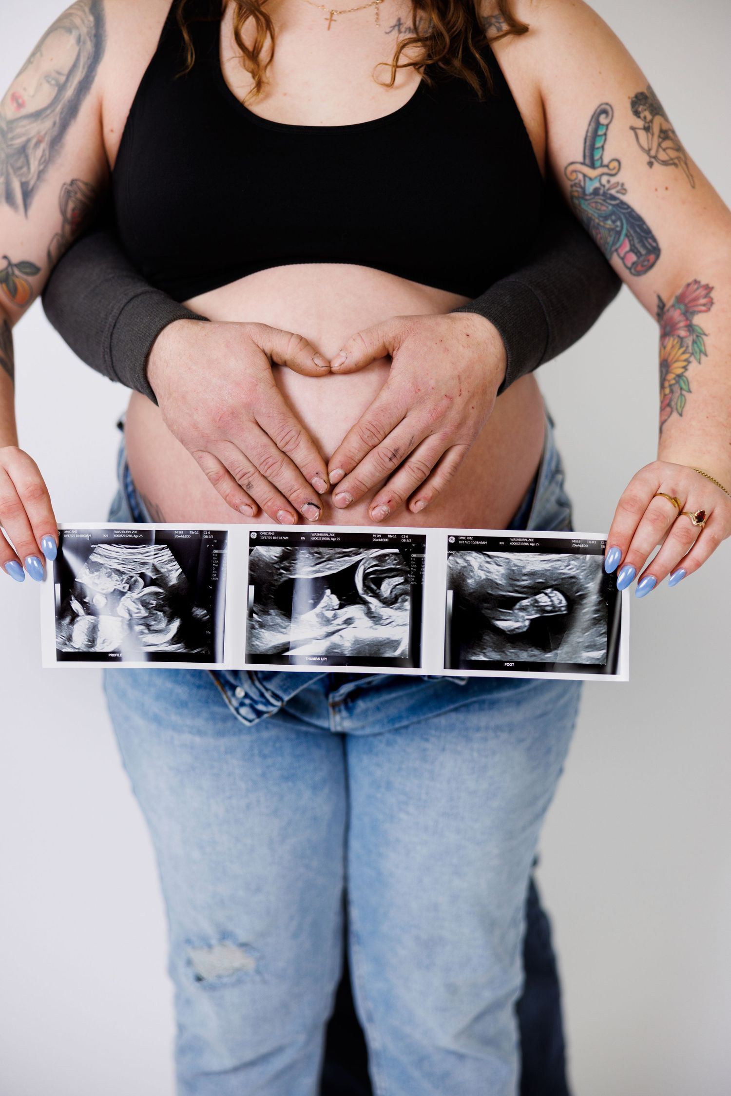 Person in jeans and black top holds ultrasound photos while showing off tattoos on their arms.
