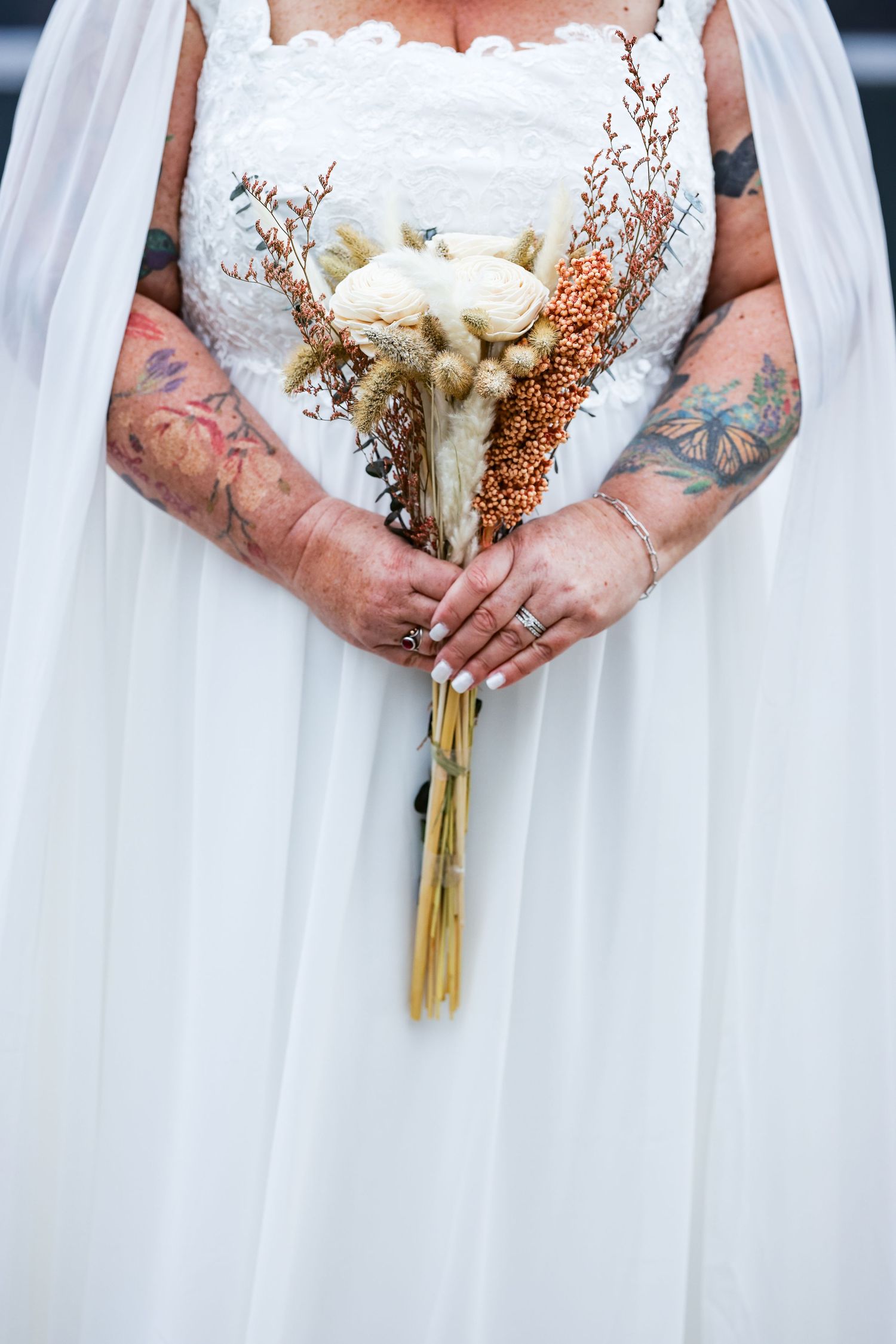 Tattooed hands holding an elegant white bouquet against a flowing white dress.