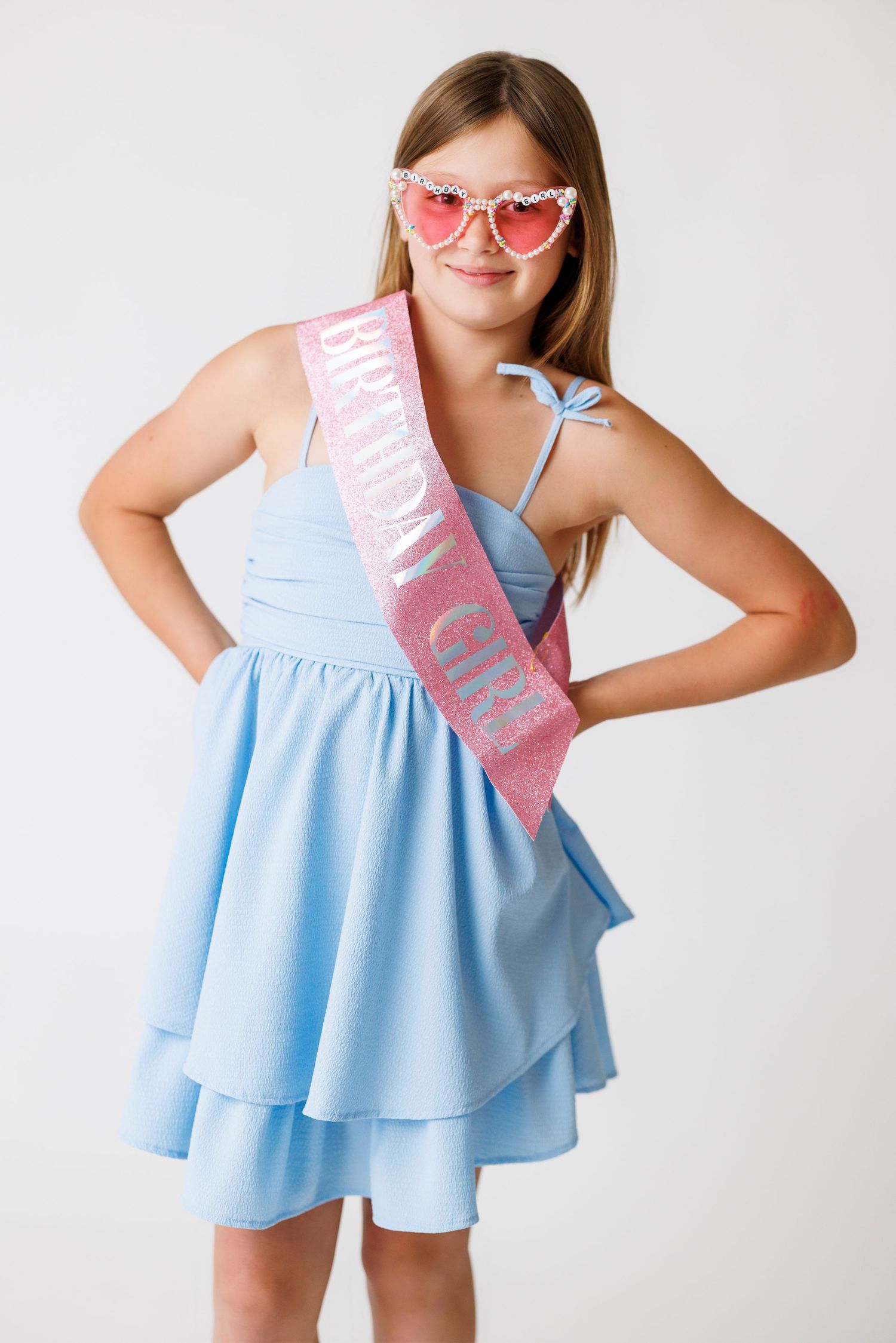 Young girl in blue dress wearing a pink Birthday Girl sash and heart-shaped sunglasses, posing confidently.