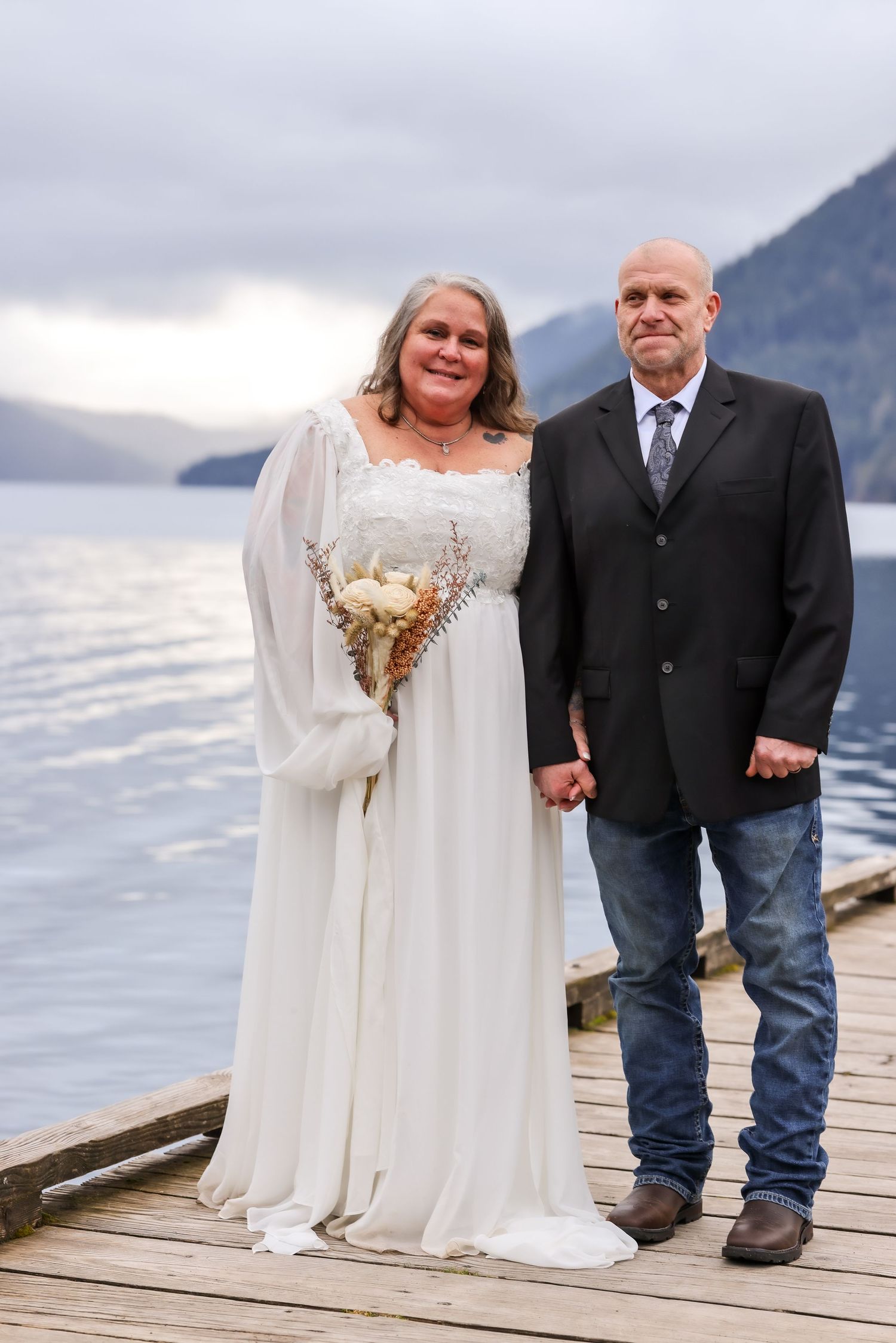 Wedding couple poses on lakeside dock with dramatic mountain scenery in background.