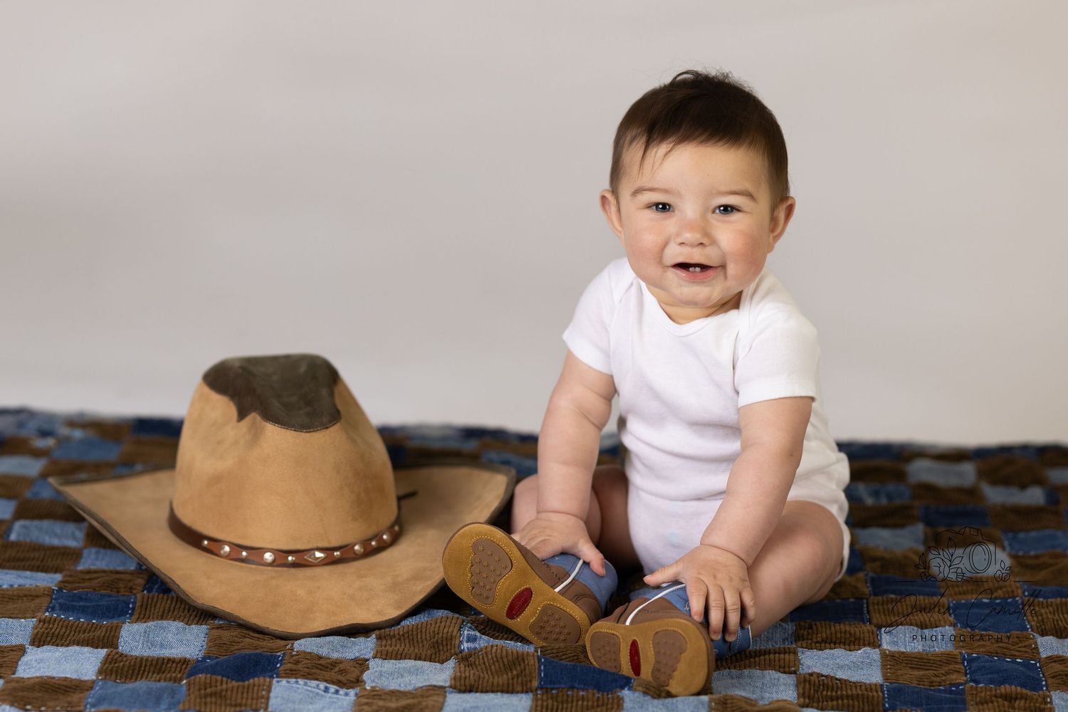 A baby in white onesie sits on a patterned rug beside a tan cowboy hat.