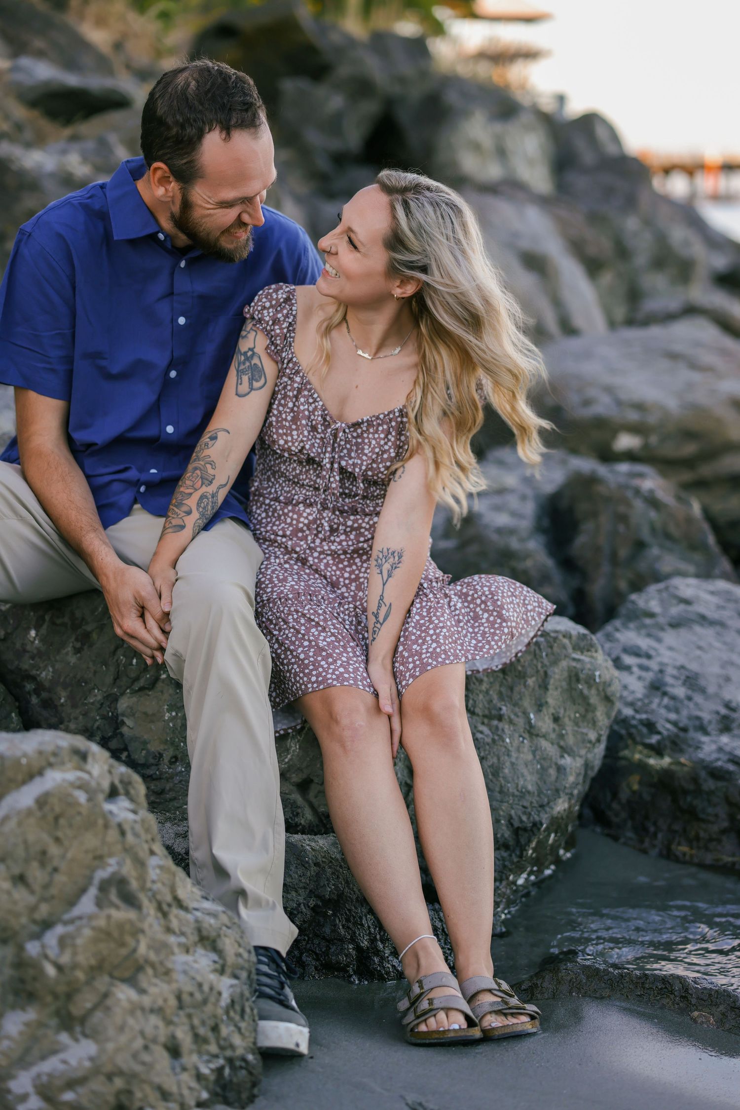 Couple sitting together on rocky beach during sunset photoshoot at ocean's edge.