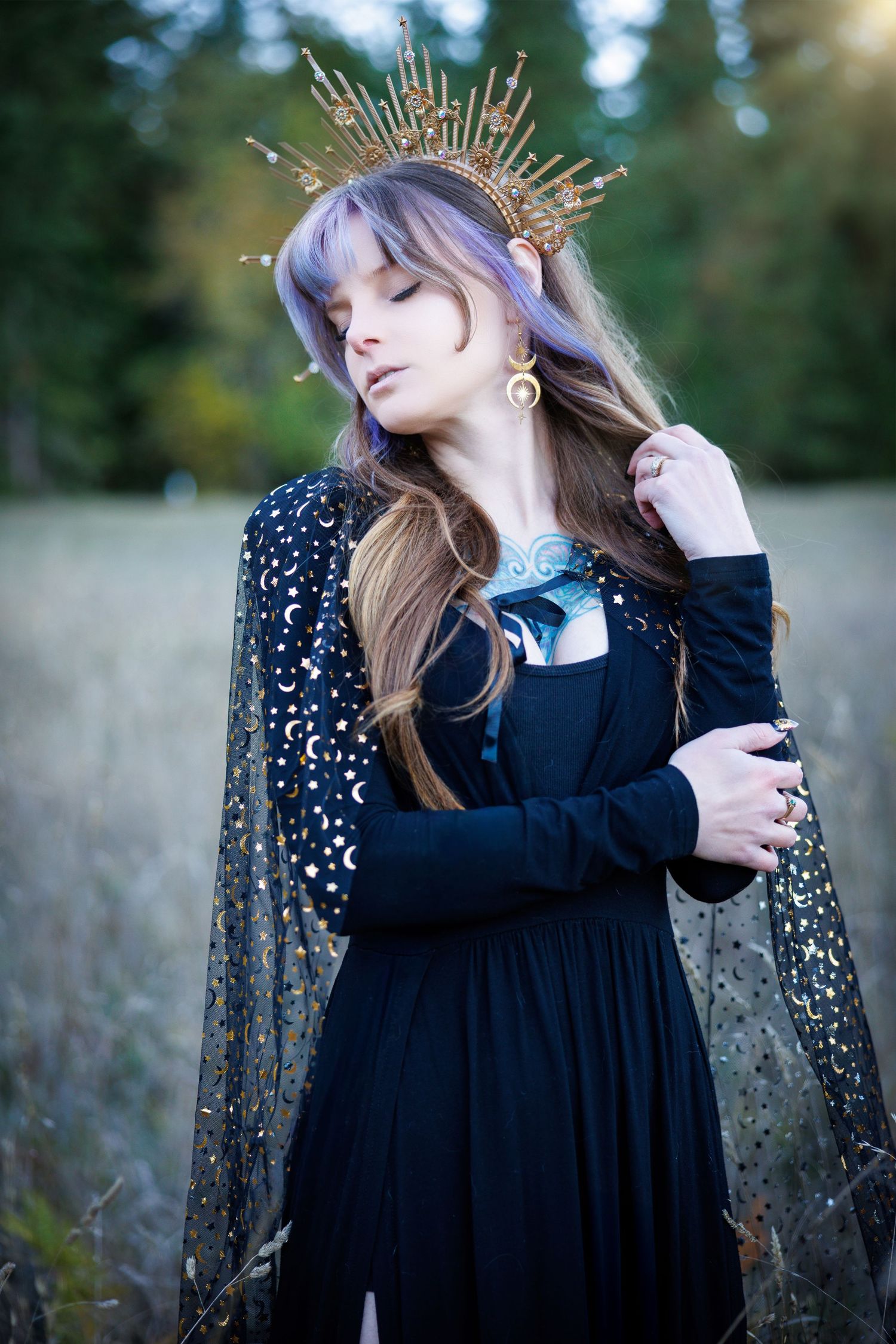 A person in a black dress and sheer cape wears a golden spiked crown while posing in a dreamy outdoor setting.