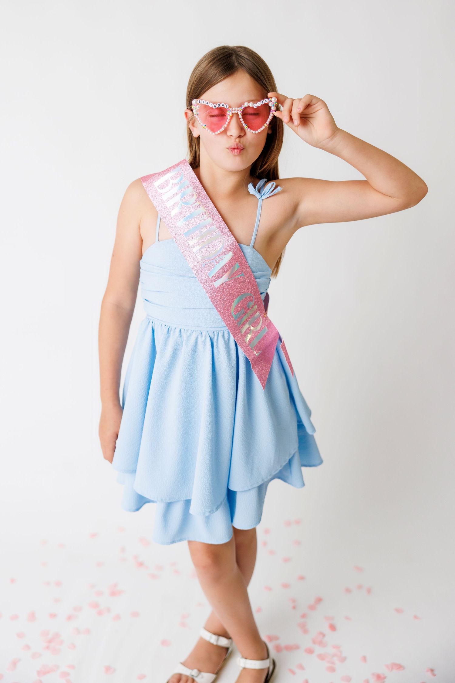 Young girl in blue dress with pink birthday sash and heart sunglasses posing on white background.
