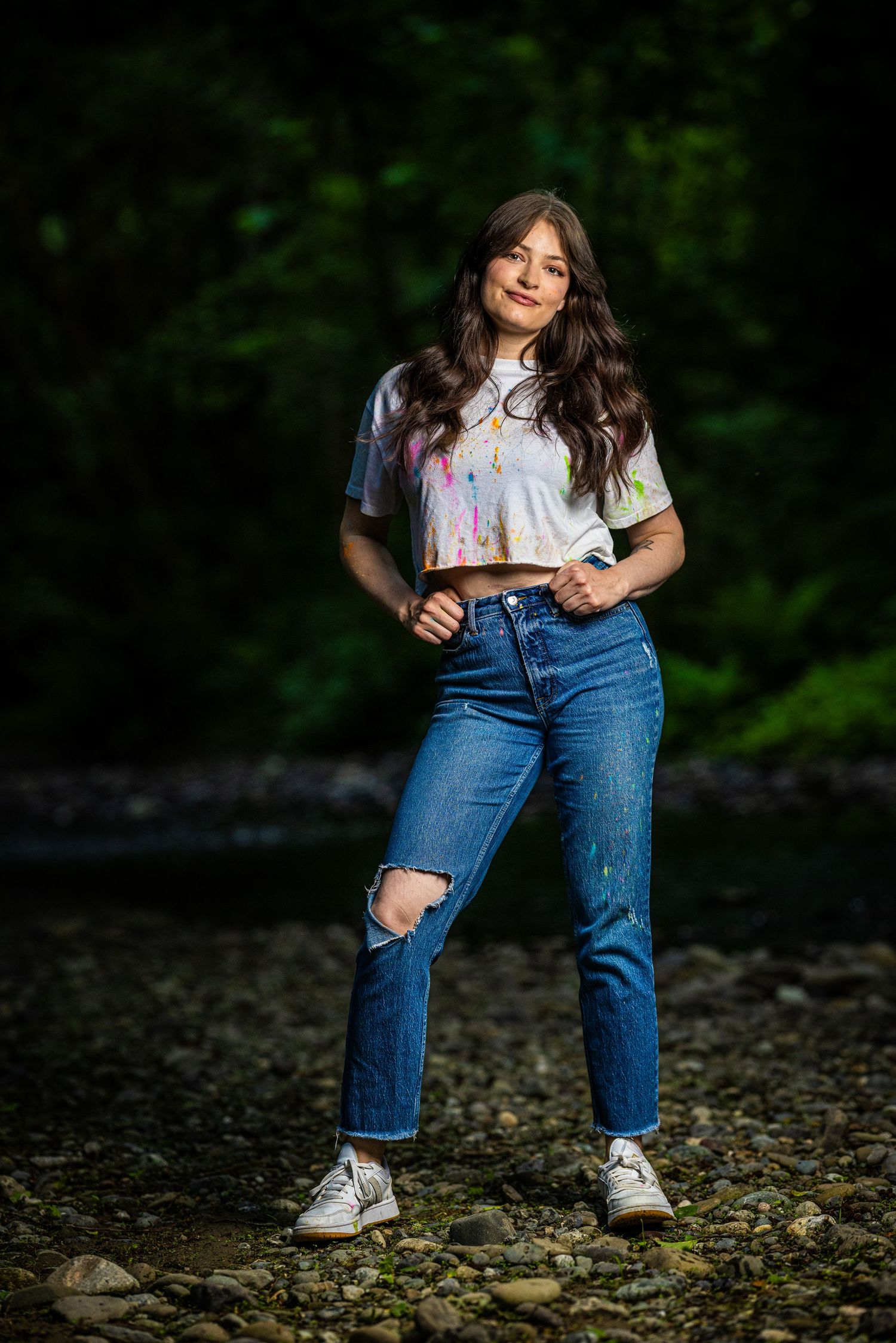 A casual outdoor portrait featuring ripped jeans and white sneakers against a dark forest background.