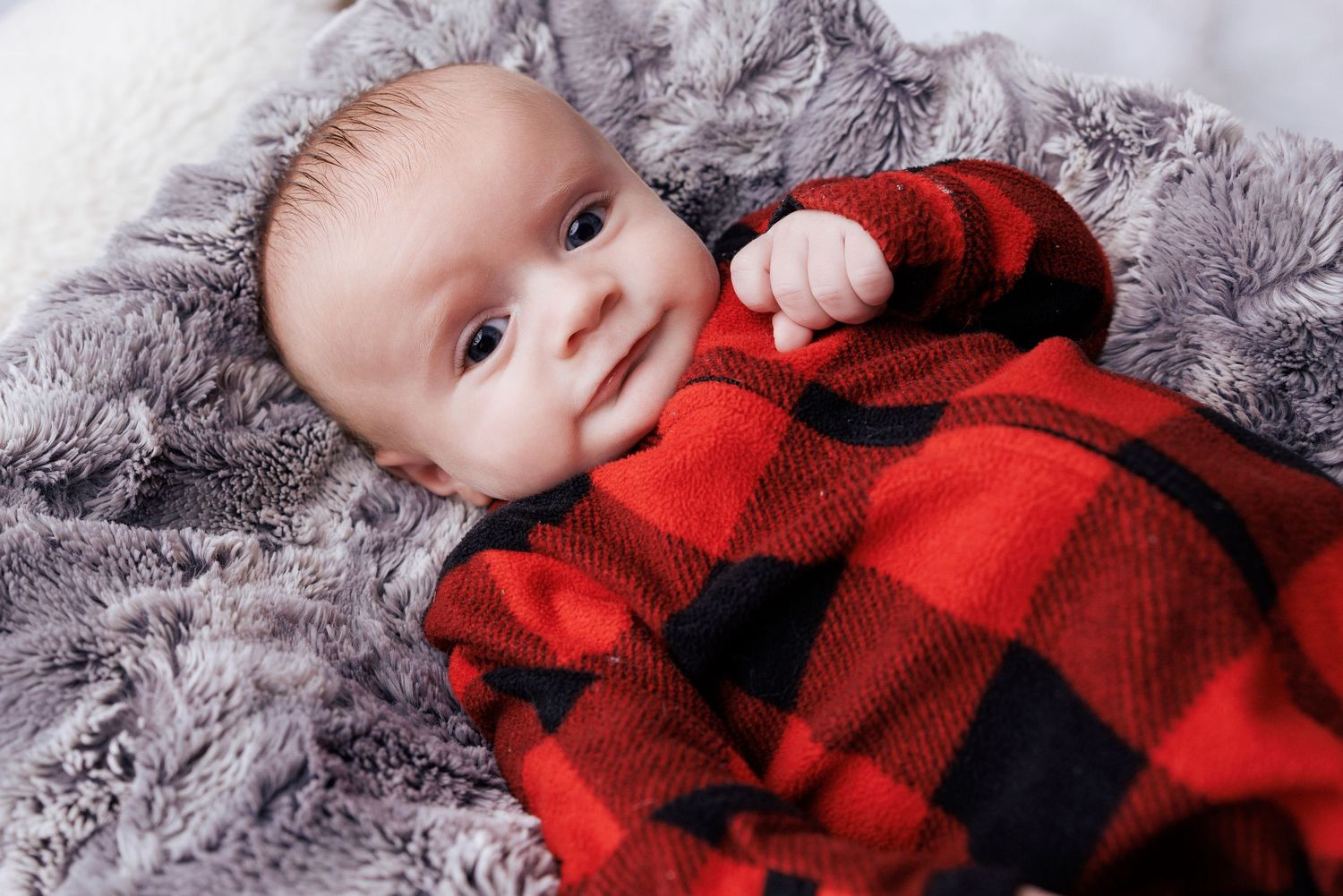 A small baby wearing a cozy red and black buffalo plaid outfit lies on a soft gray blanket.