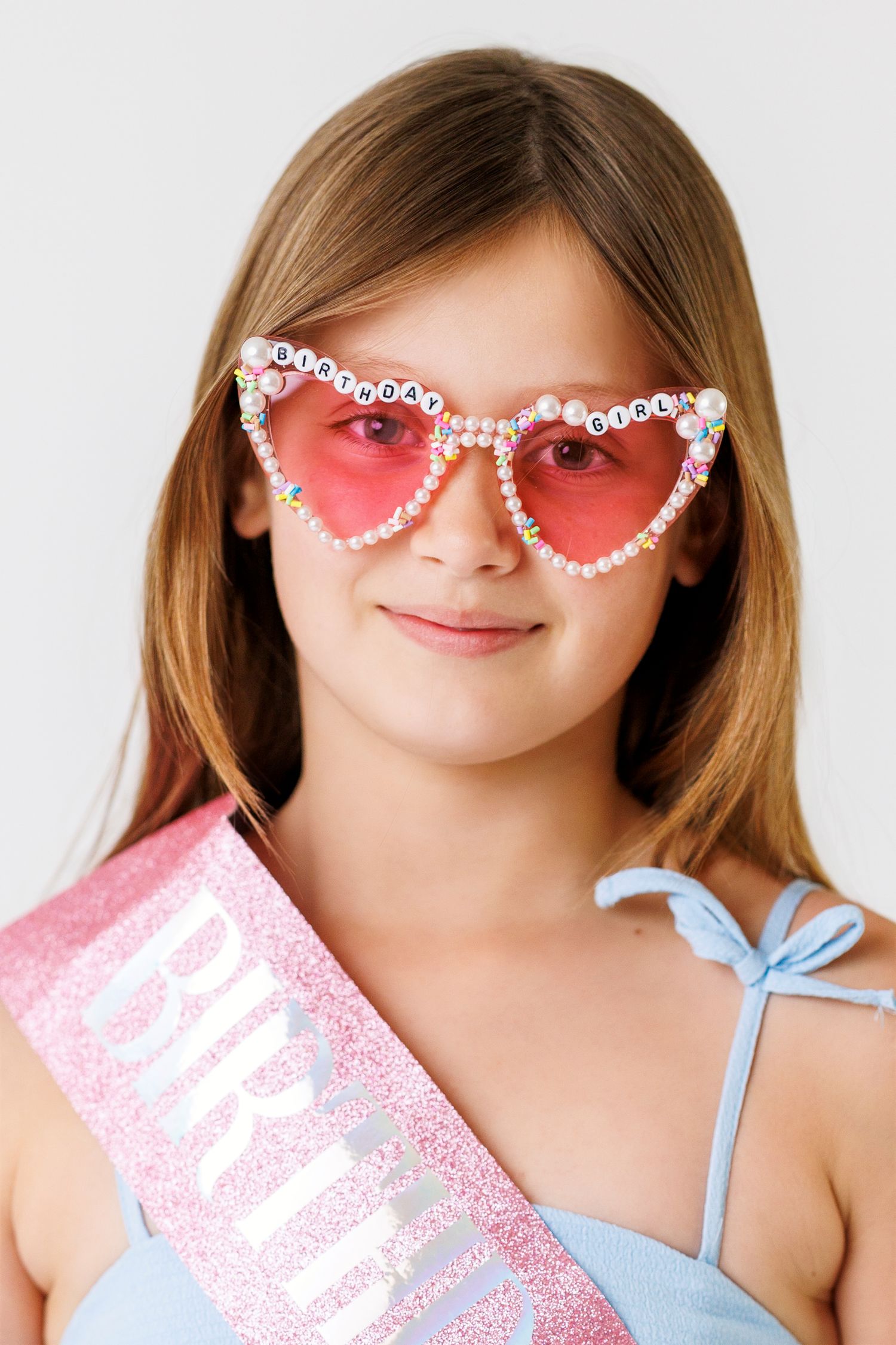 Young girl wearing pink heart-shaped Birthday Girl glasses and glittery pink sash, smiling at camera.