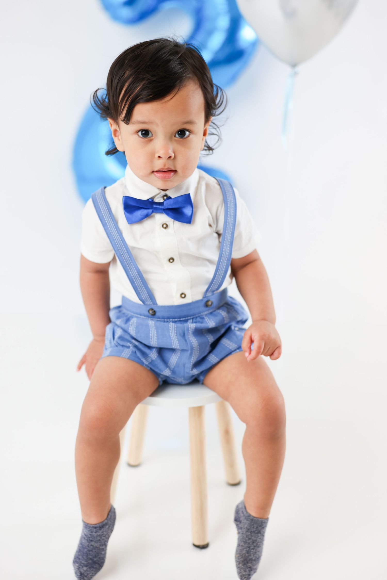 Adorable baby boy in blue suspenders, bow tie, and white shirt sits on a stool with blue balloons.
