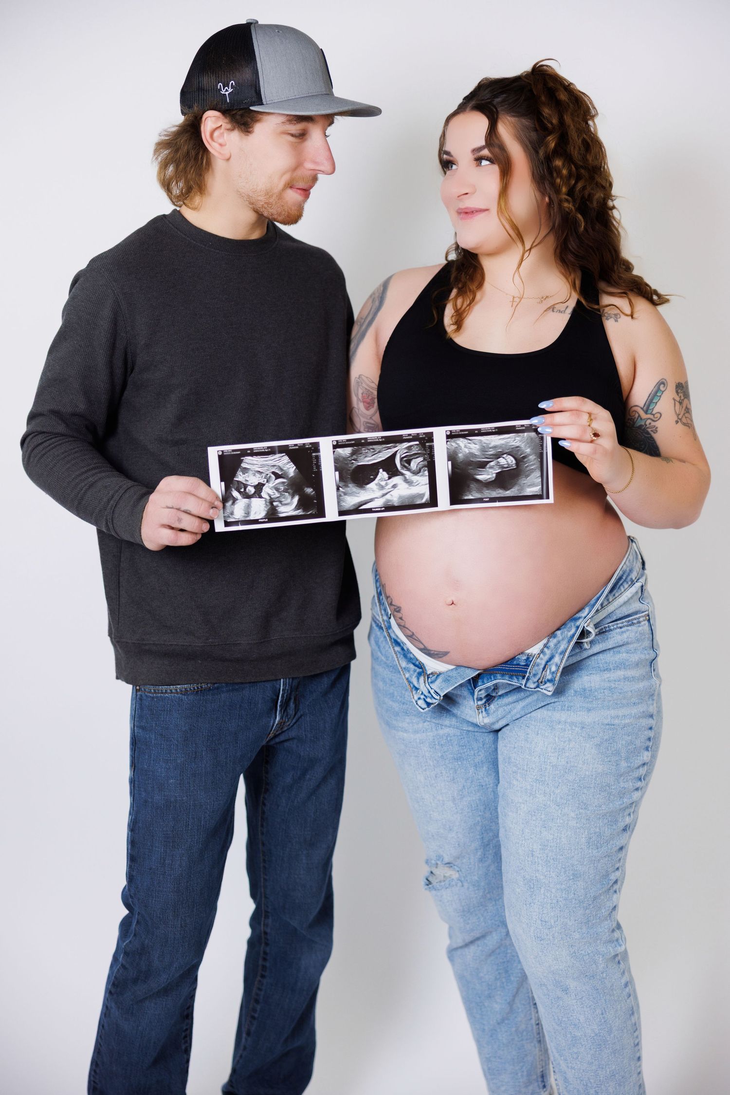 A couple poses for a pregnancy photo shoot with ultrasound images against a white backdrop.
