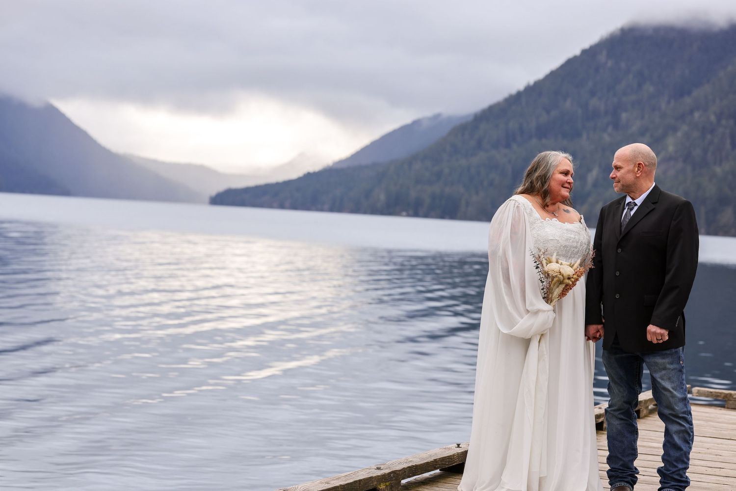 A couple stands together against a scenic mountain lake backdrop during their wedding photoshoot.