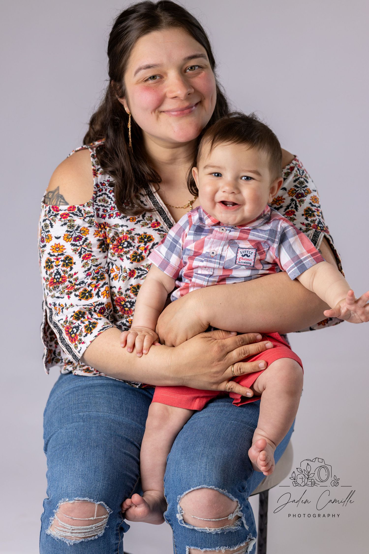 A mother in ripped jeans and floral top shares joyful moments with her baby in a plaid shirt against a gray backdrop.
