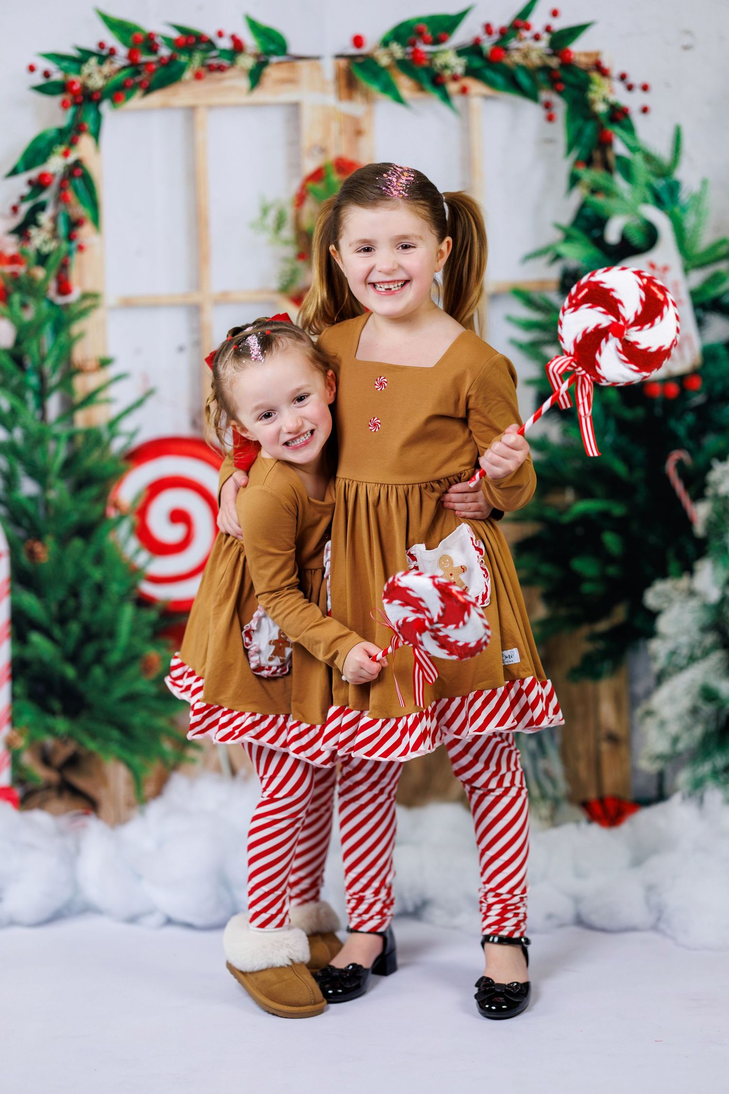 Two children in matching brown dresses and candy cane striped leggings pose with Christmas decorations in a festive setting.