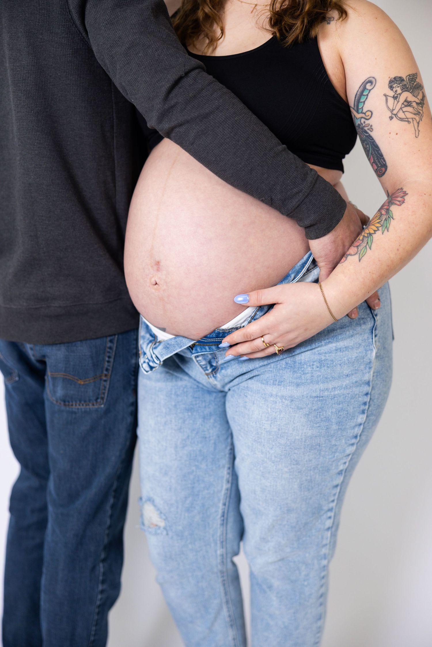 Close-up of pregnant belly with partner's hand resting on it, woman wearing black tank top and light denim.