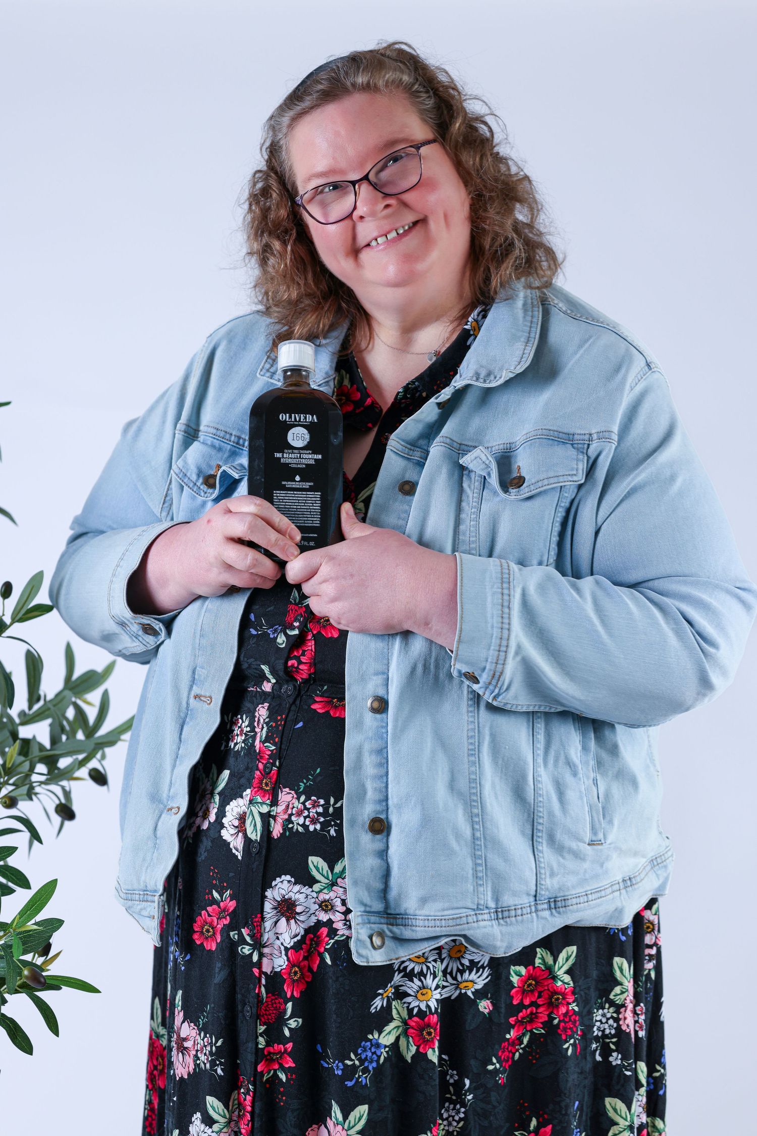 Photographer in denim jacket and floral dress posing with camera against white background.