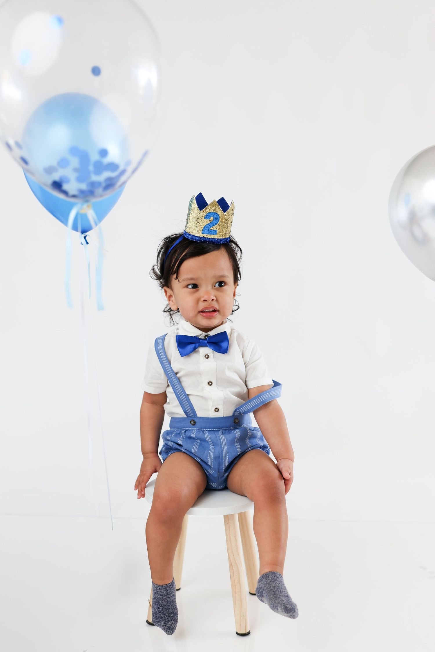 Toddler boy in blue suspenders and bow tie wearing a crown, sitting on chair with balloons for 2nd birthday.