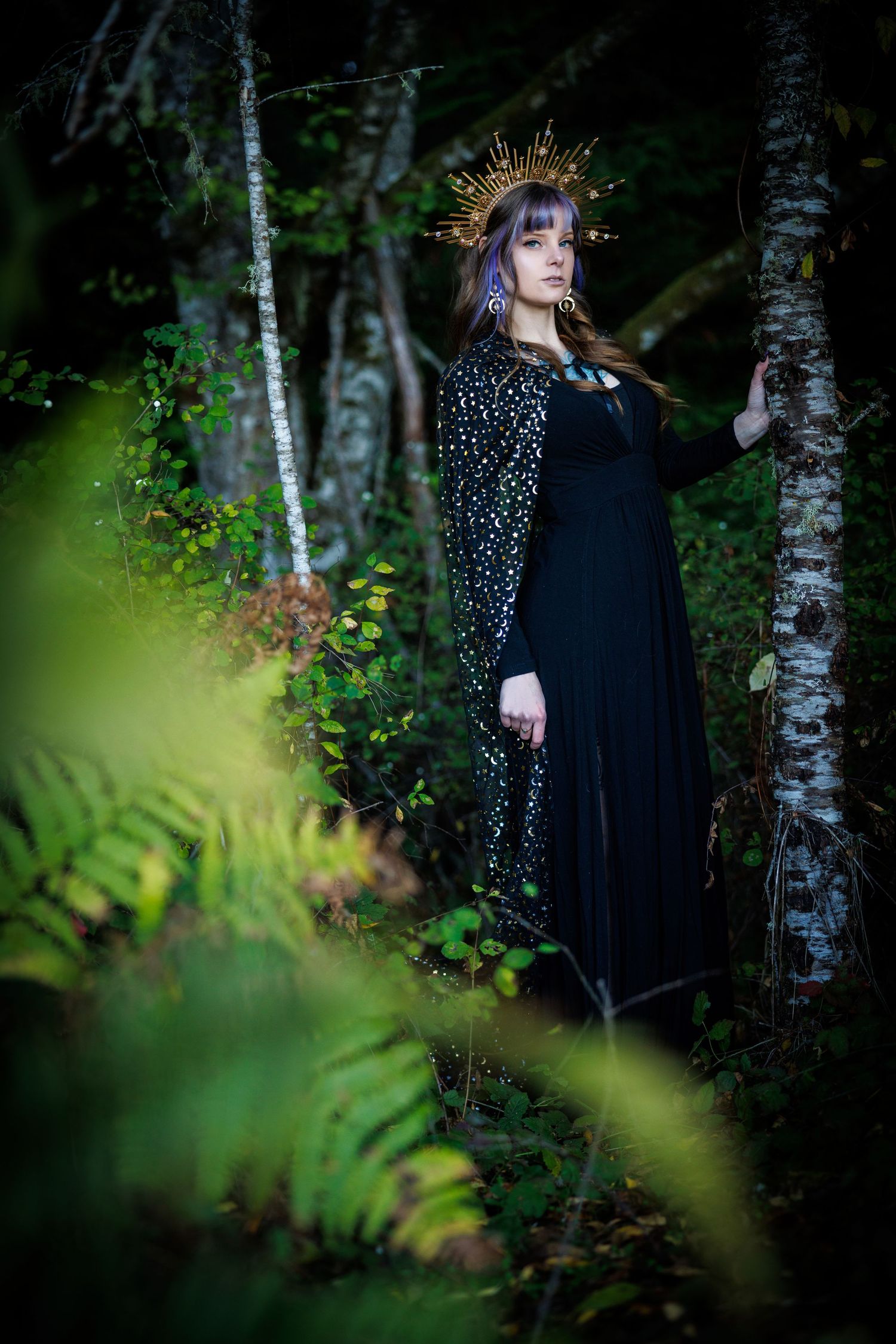 A figure in a black dress and ornate crown stands mysteriously between birch trees in a misty forest setting.