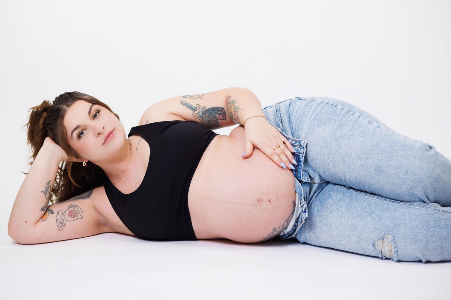 A pregnant person in a black tank top and jeans lies on their side on a white background.