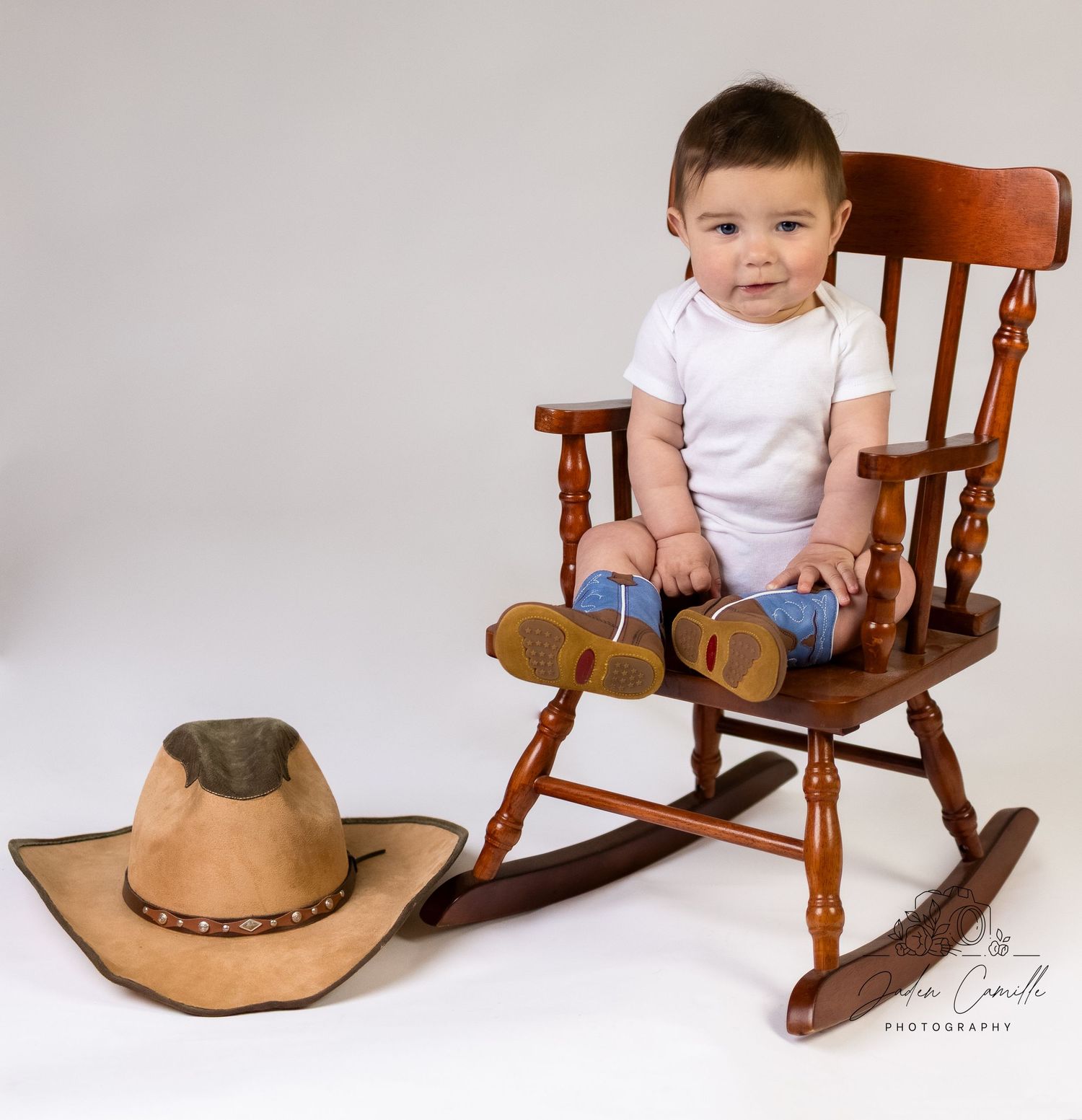 A small child sits in a wooden rocking chair next to a brown cowboy hat in a studio setting.
