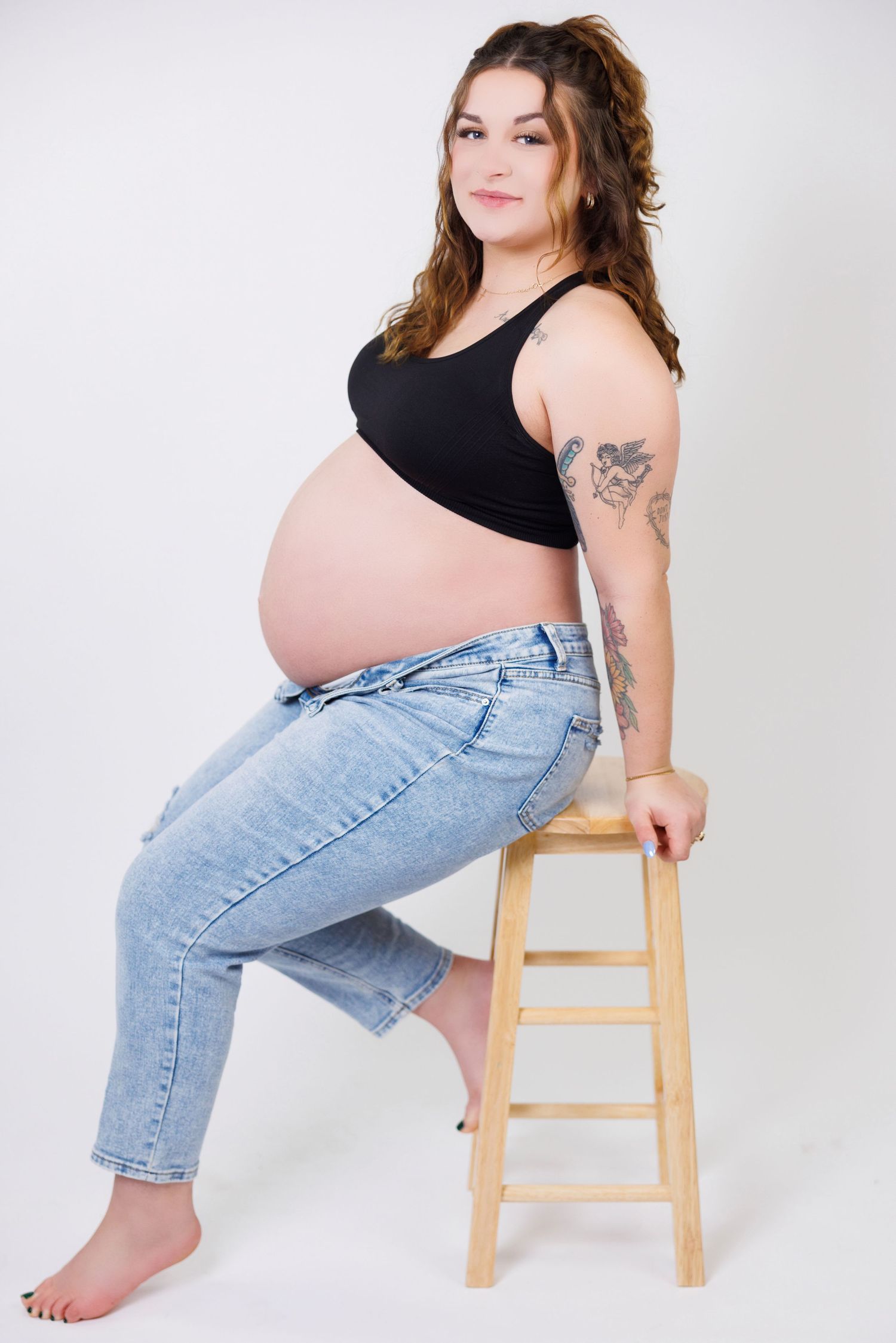 A series of maternity photos showing someone in jeans and black top posing casually on a wooden stool against white backdrop.