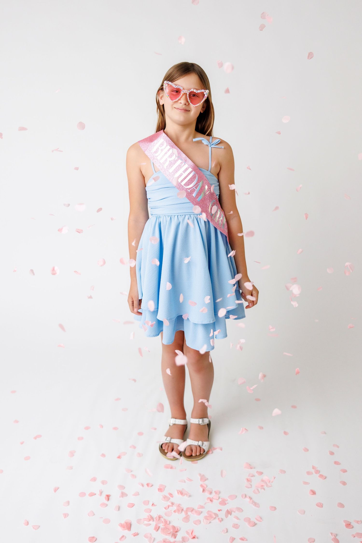 A young girl in a blue dress with a pink birthday sash and heart sunglasses, surrounded by falling confetti.