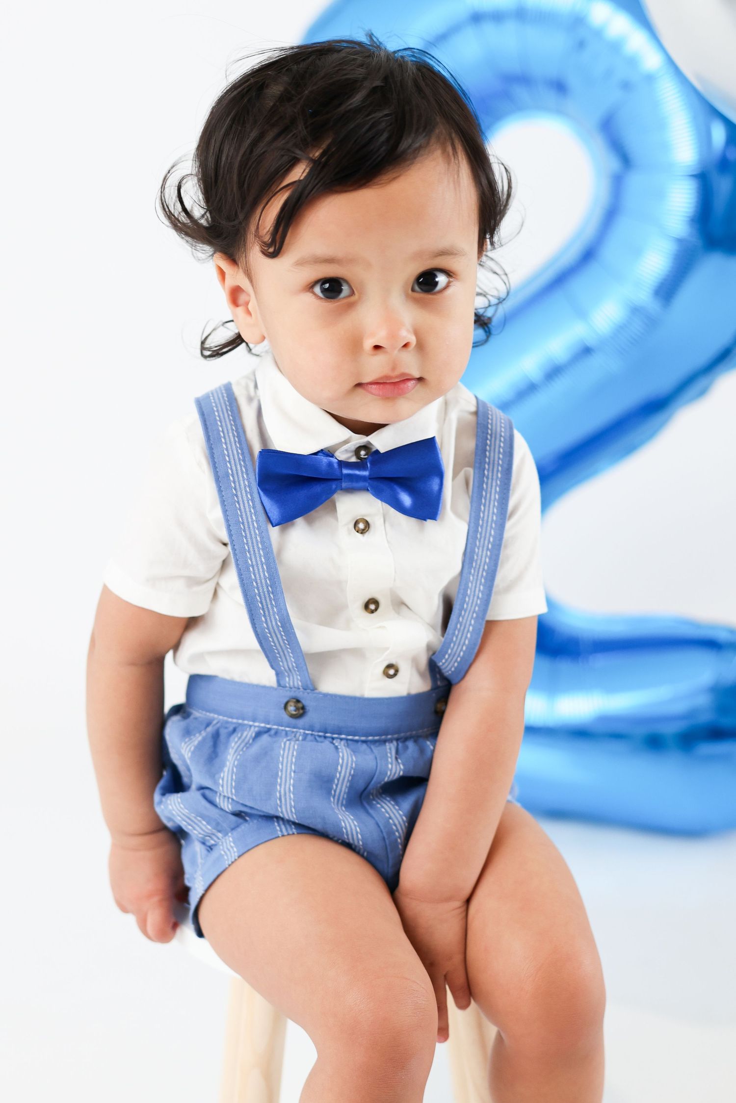 Adorable toddler boy in blue bow tie and suspenders outfit sitting near a number 2 balloon for birthday photo.