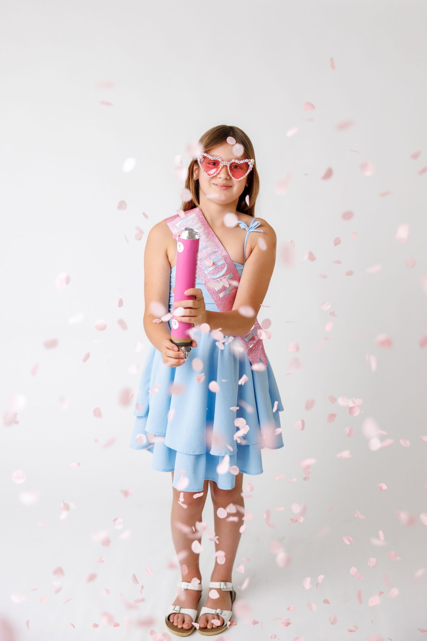 Young girl in blue dress and pink heart sunglasses holding confetti cannon with pink confetti falling around her.