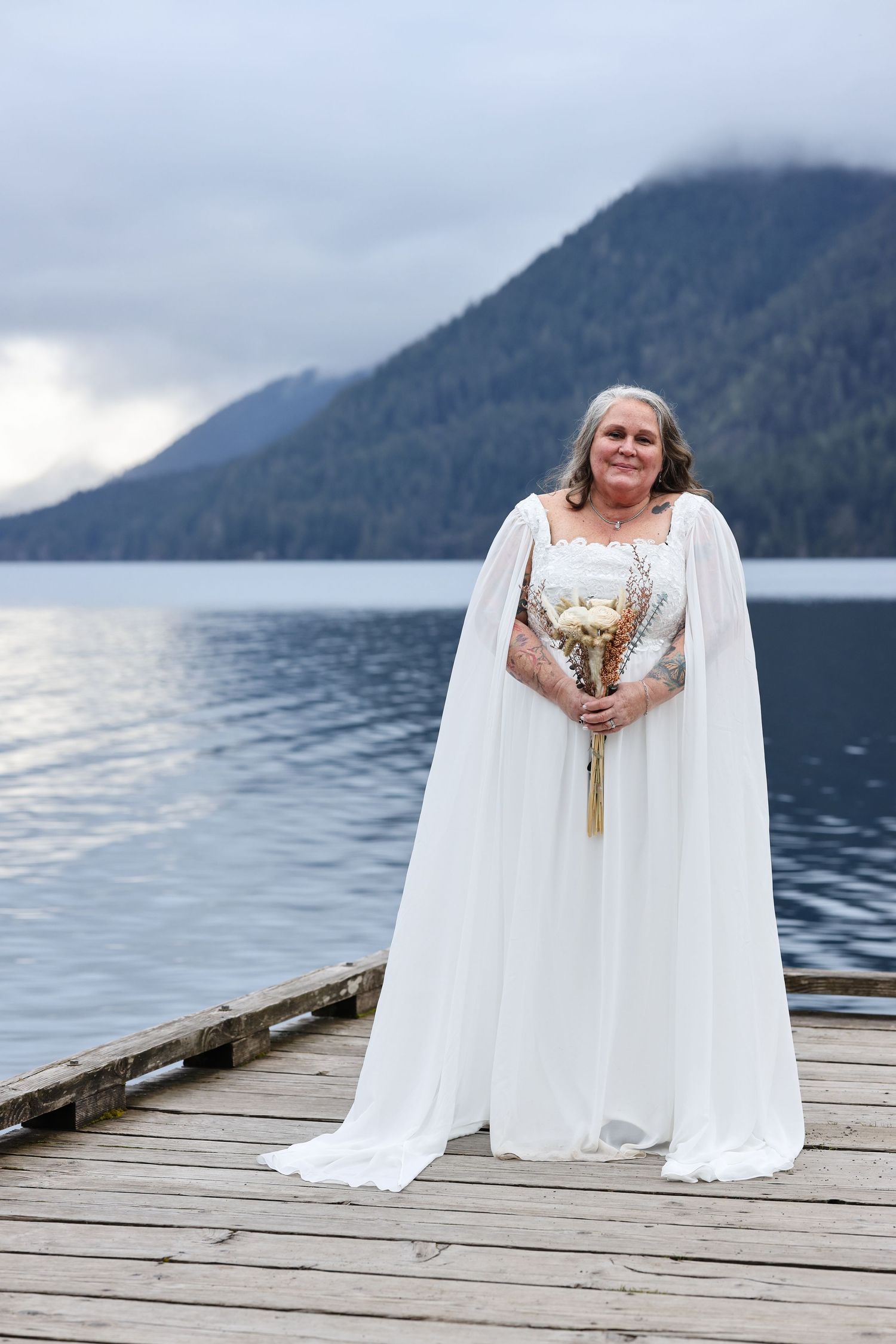 Bride in flowing white dress holds bouquet while standing on dock overlooking mountain lake.